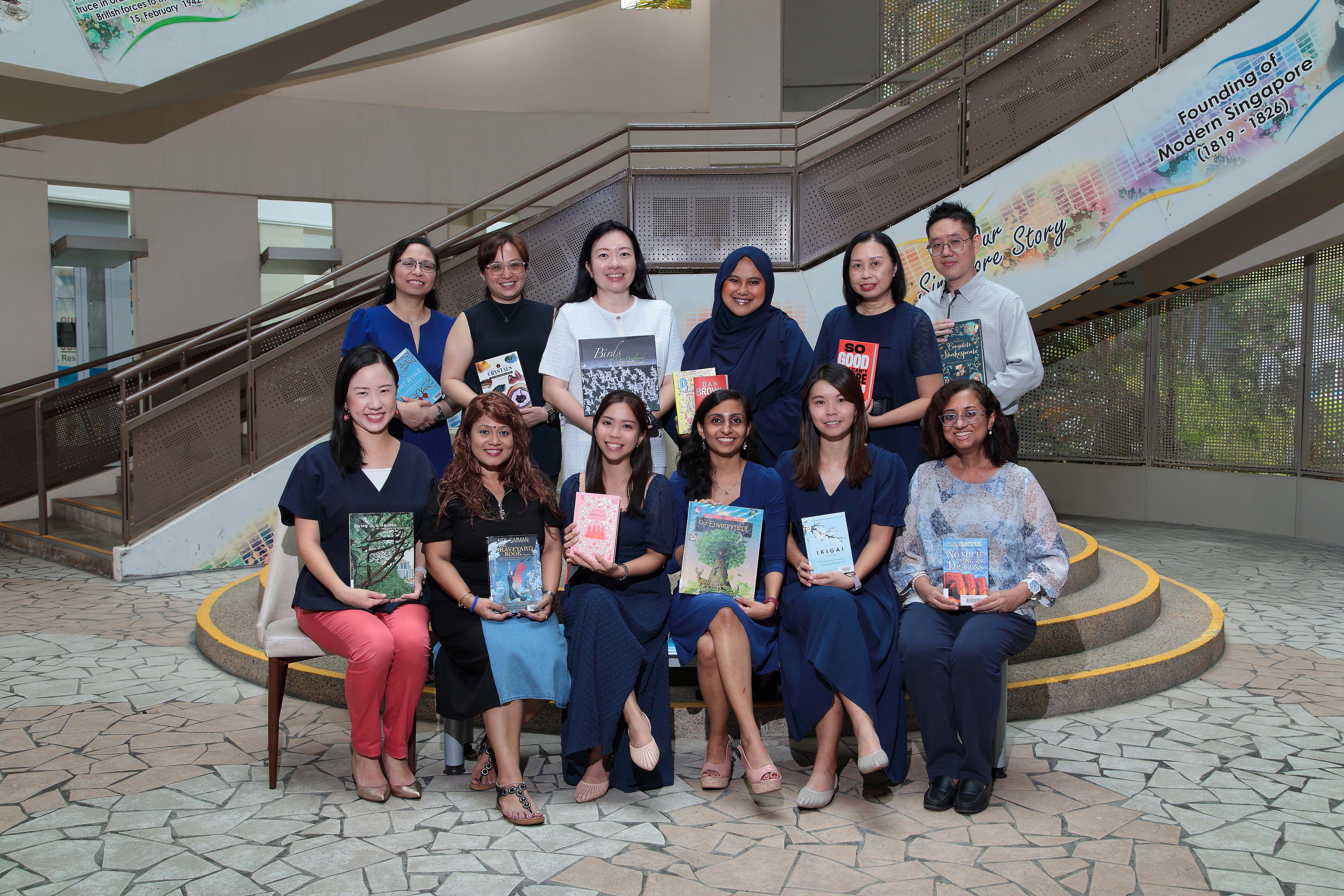 EL Department staff pose in two rows on steps, each holding a book, inside a building with Singapore history murals.