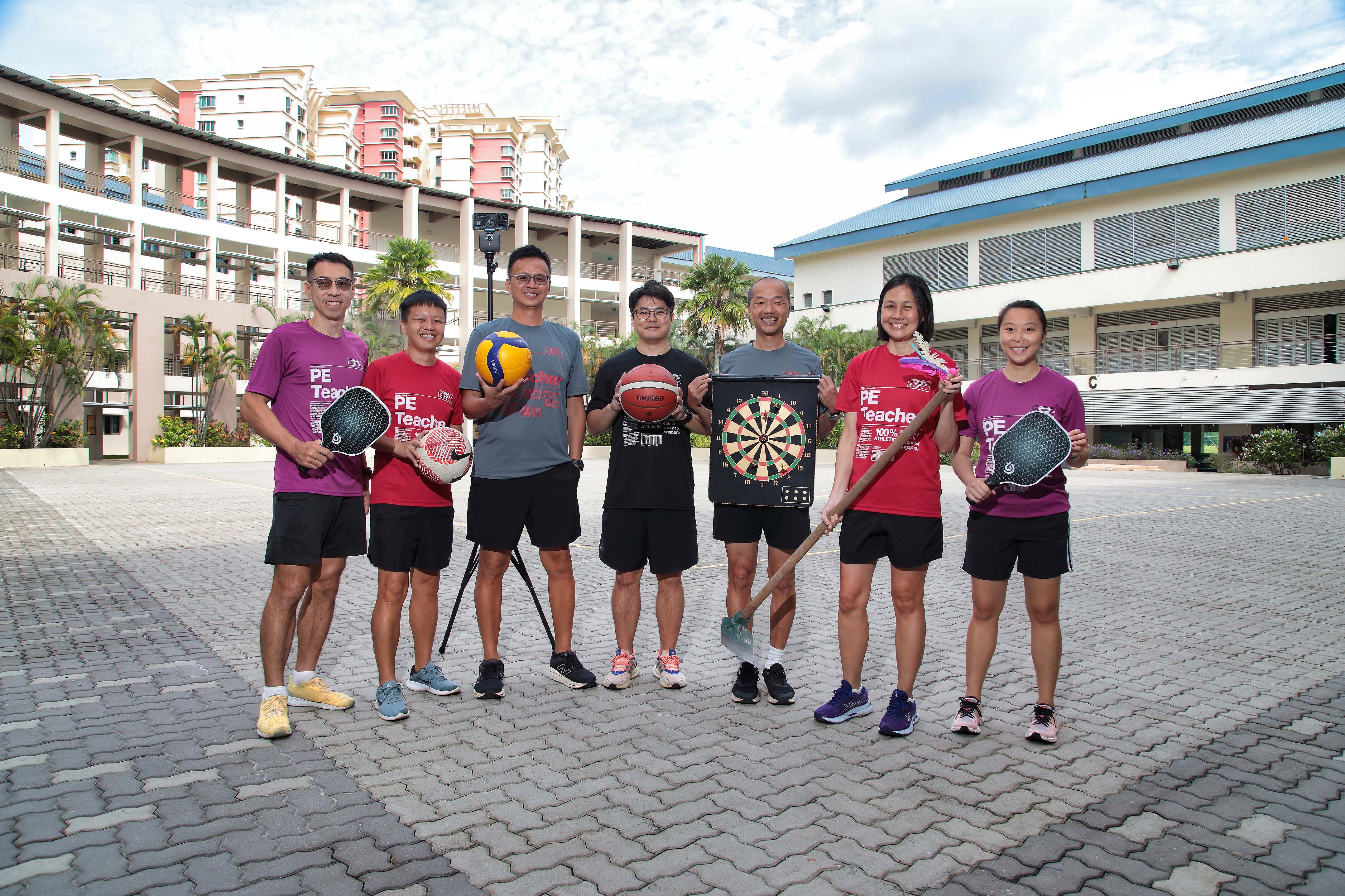 PE Department teachers pose outdoors holding sports equipment including a basketball, volleyball, dartboard, and paddles.