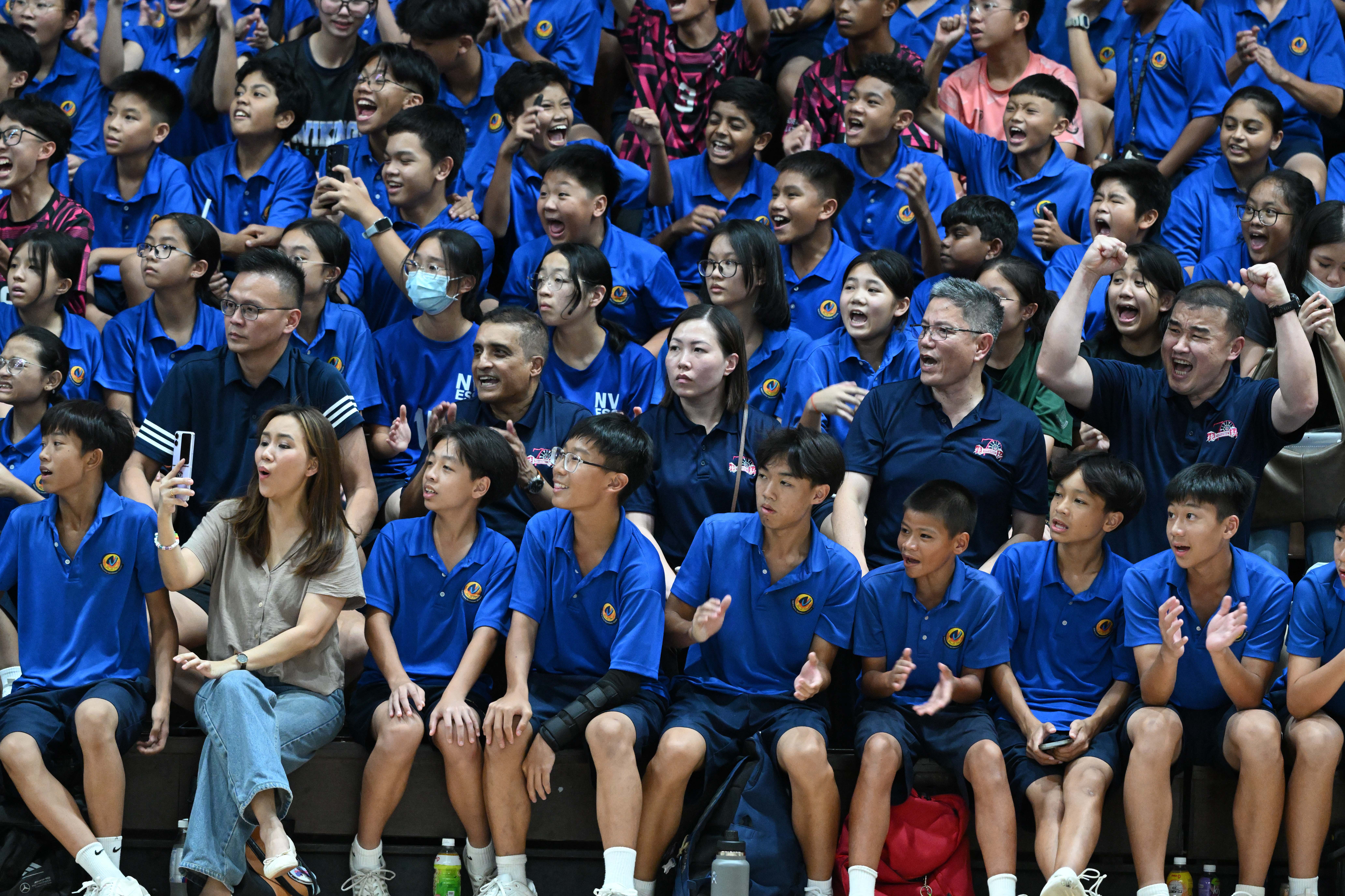 A crowd of students and school staff in blue shirts cheering enthusiastically from the indoor stadium.