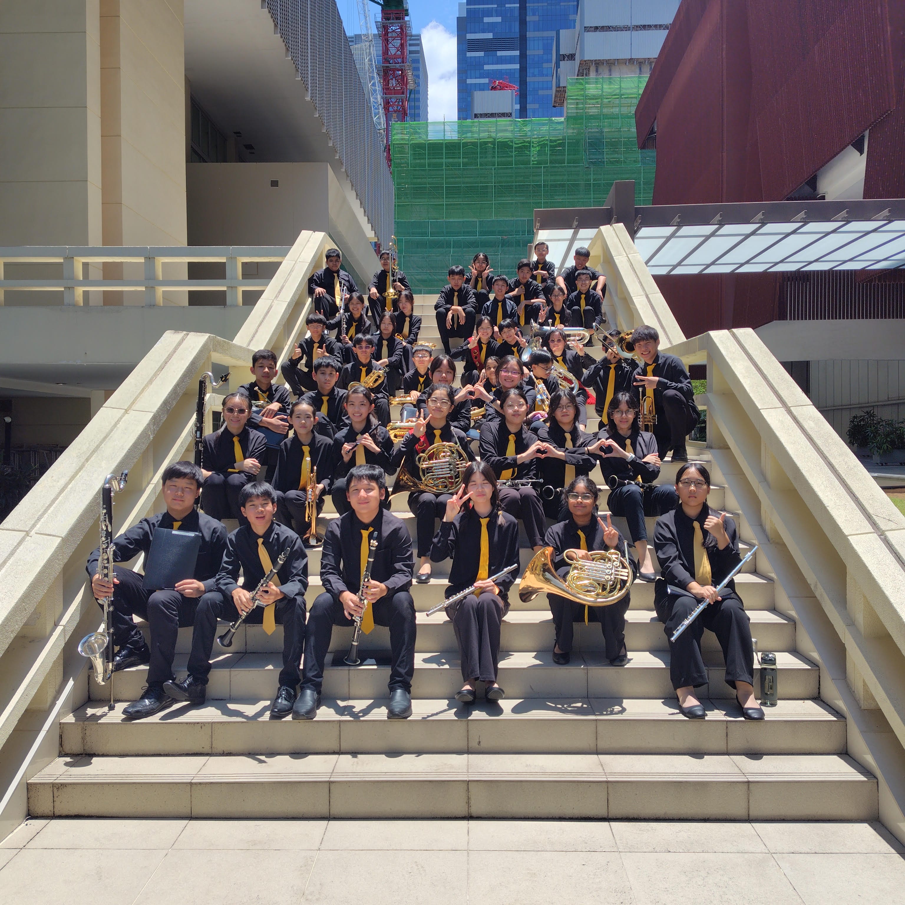 NV Symphonic Band members in black uniforms with yellow ties poses on outdoor stairs holding instruments like flutes and French horns.
