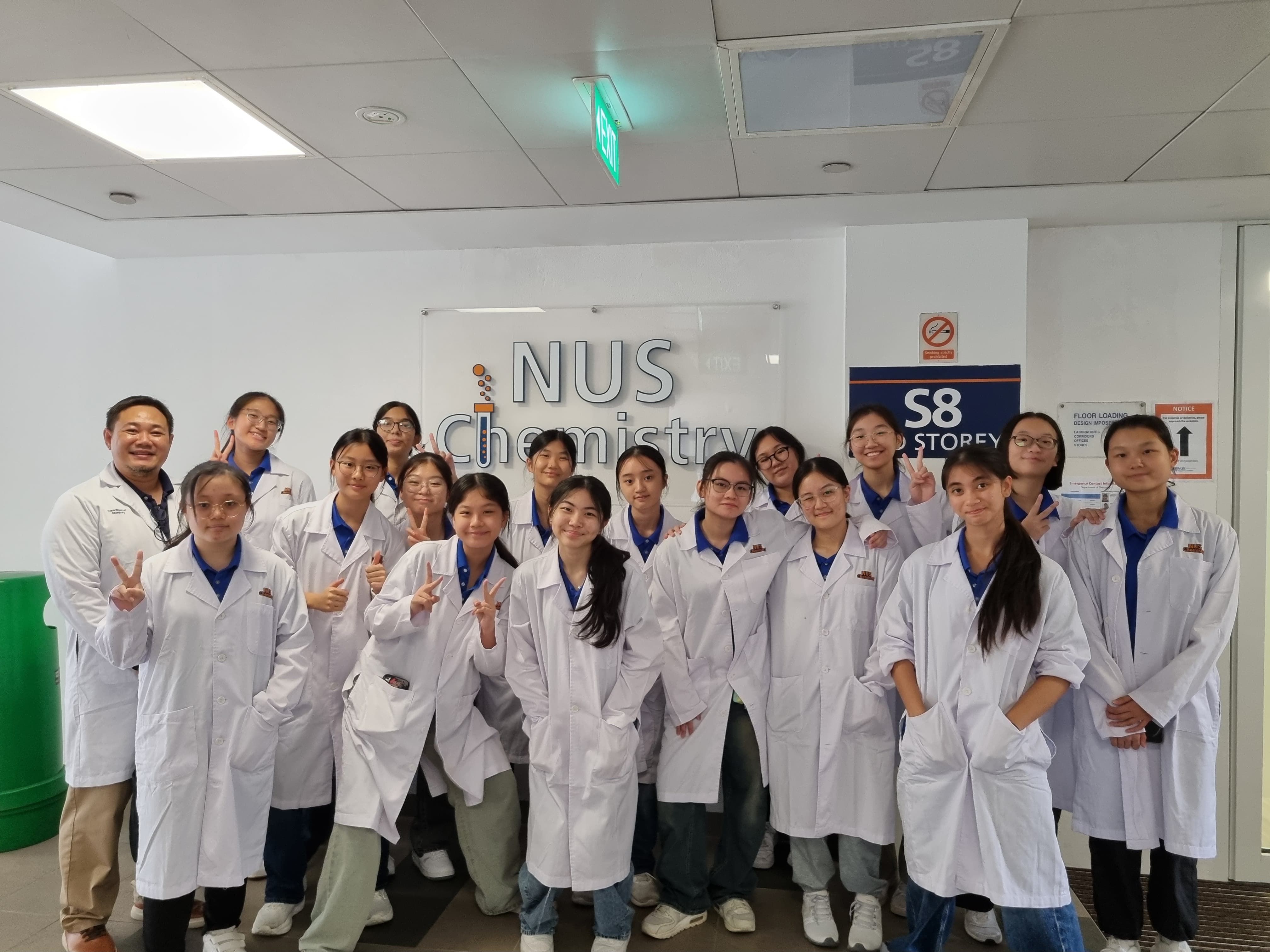 NV students and an instructor in white lab coats pose in front of the NUS Chemistry sign.