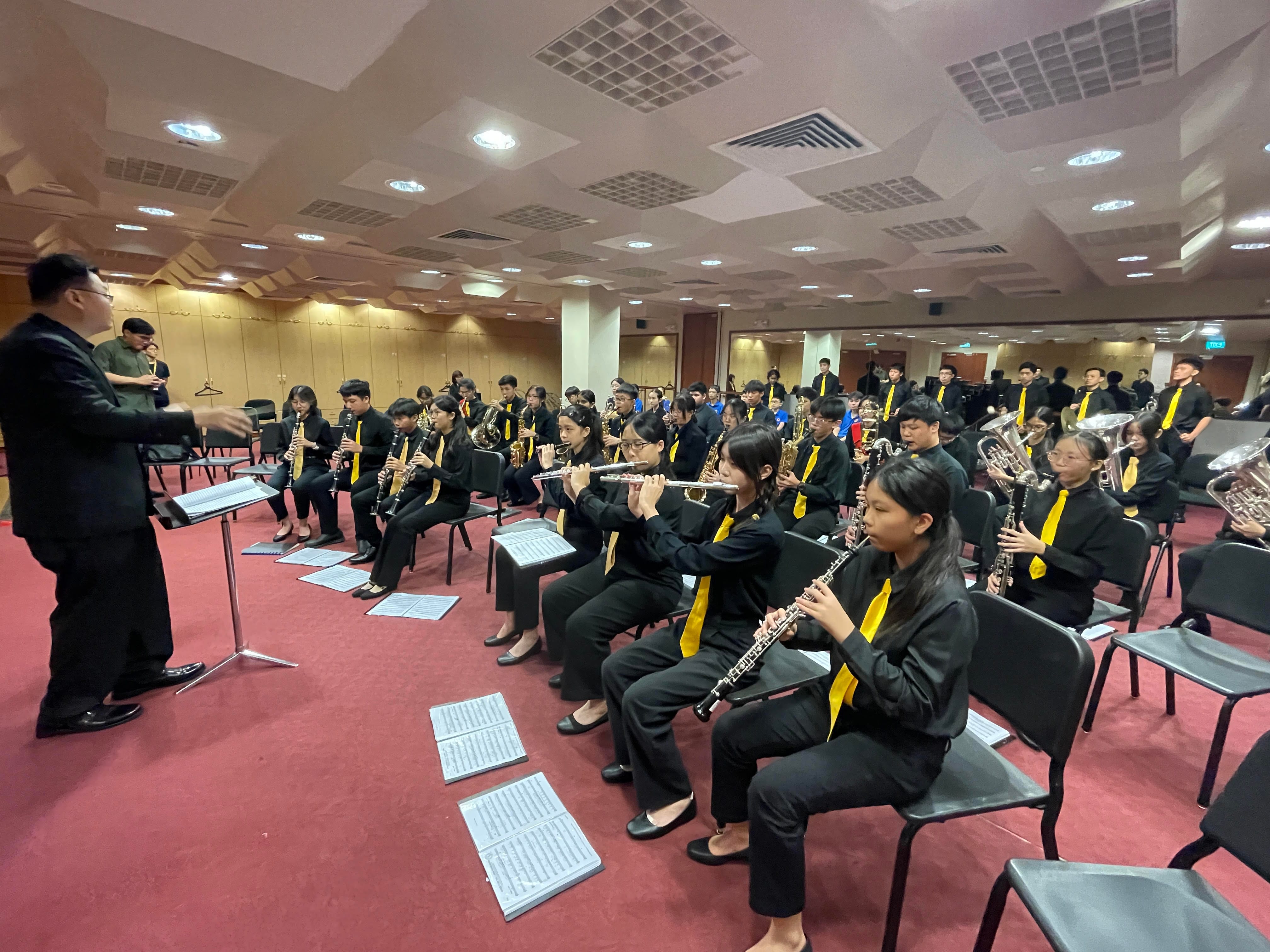 A conductor leads a youth wind band in black uniforms with yellow ties playing woodwind and brass instruments indoors.