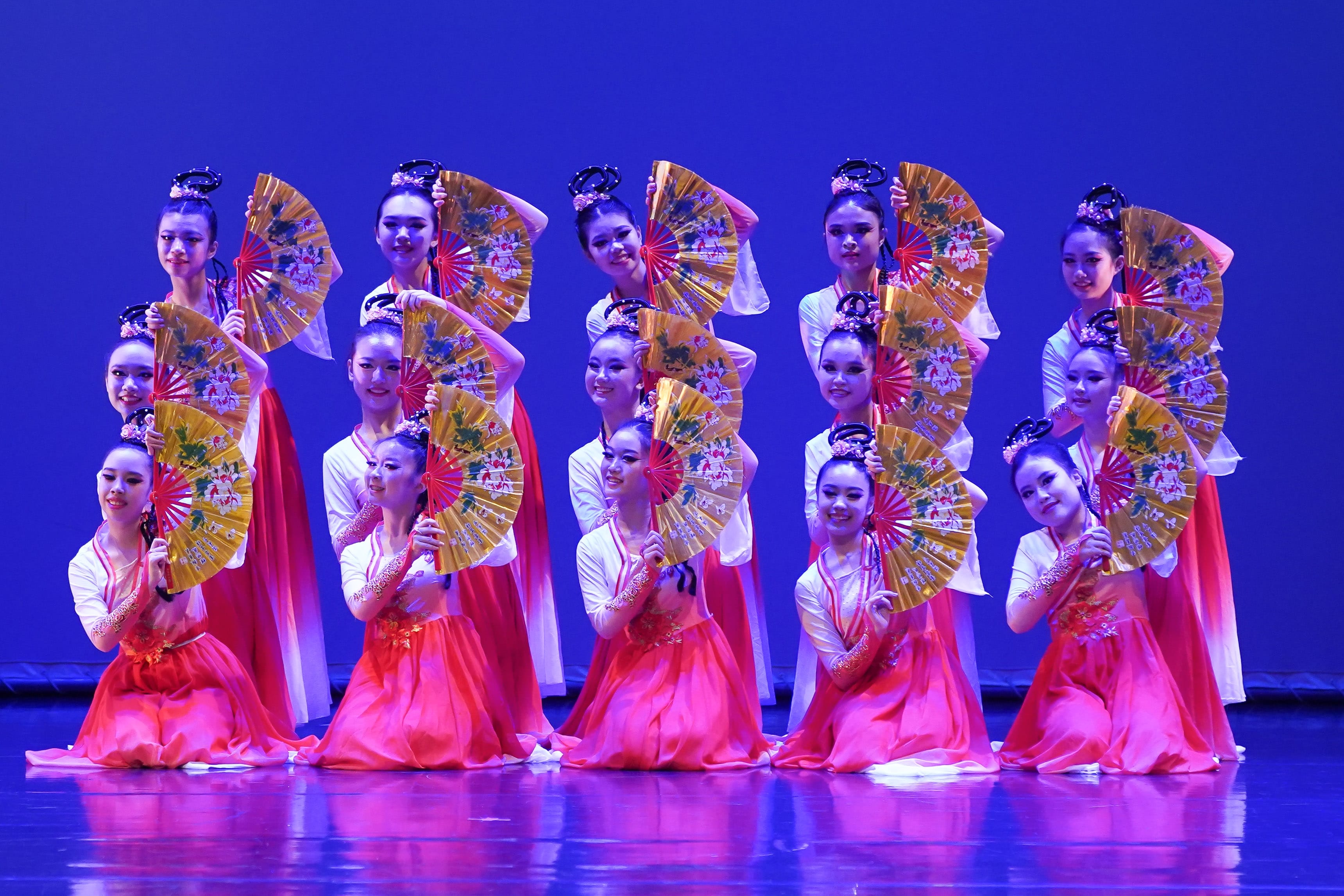Dancers in red and white traditional costumes hold golden folding fans on a blue-lit stage.