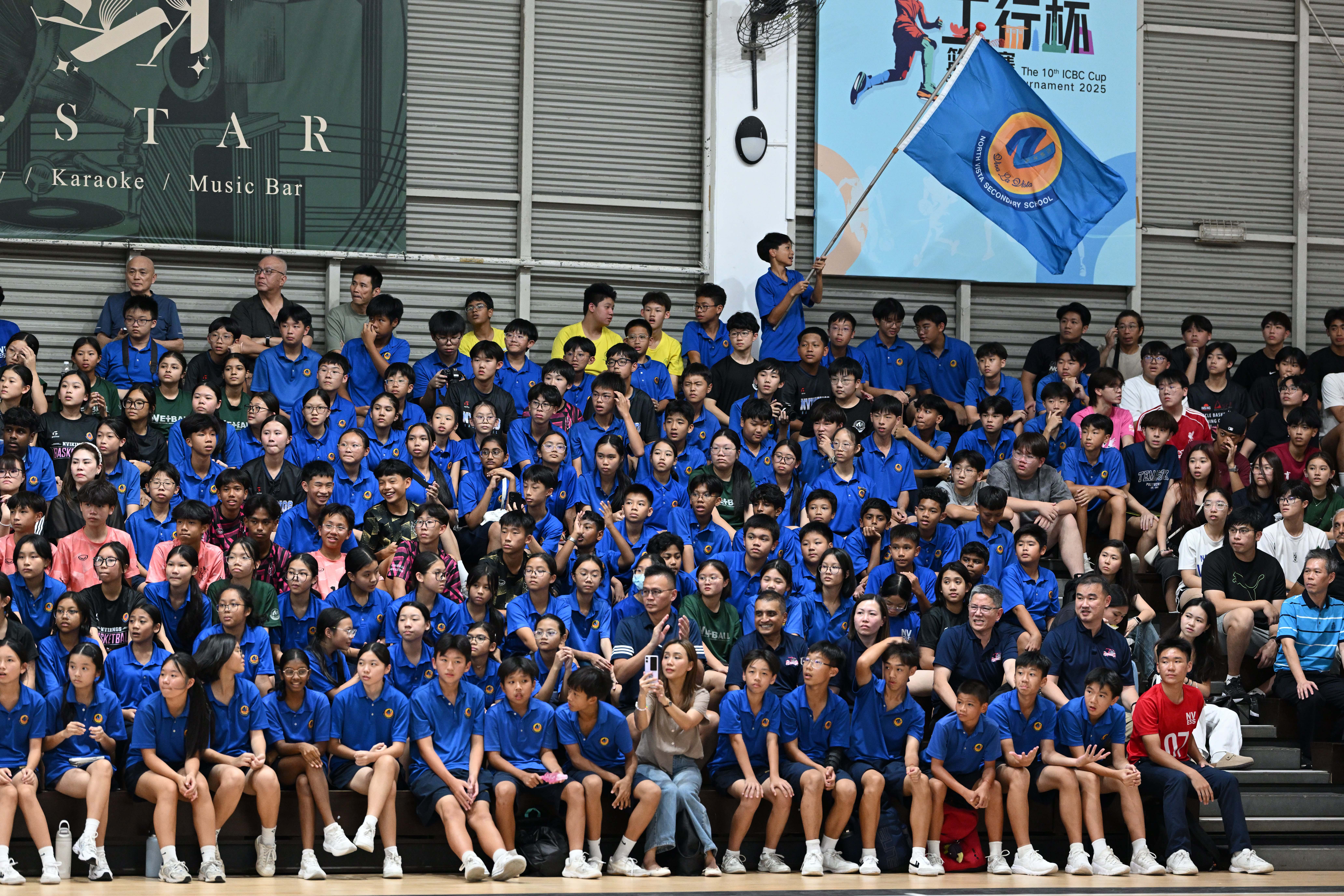 Large group of students and supporters in a sports hall, many wearing blue shirts, with one student holding a school flag high, and banners on the walls.
