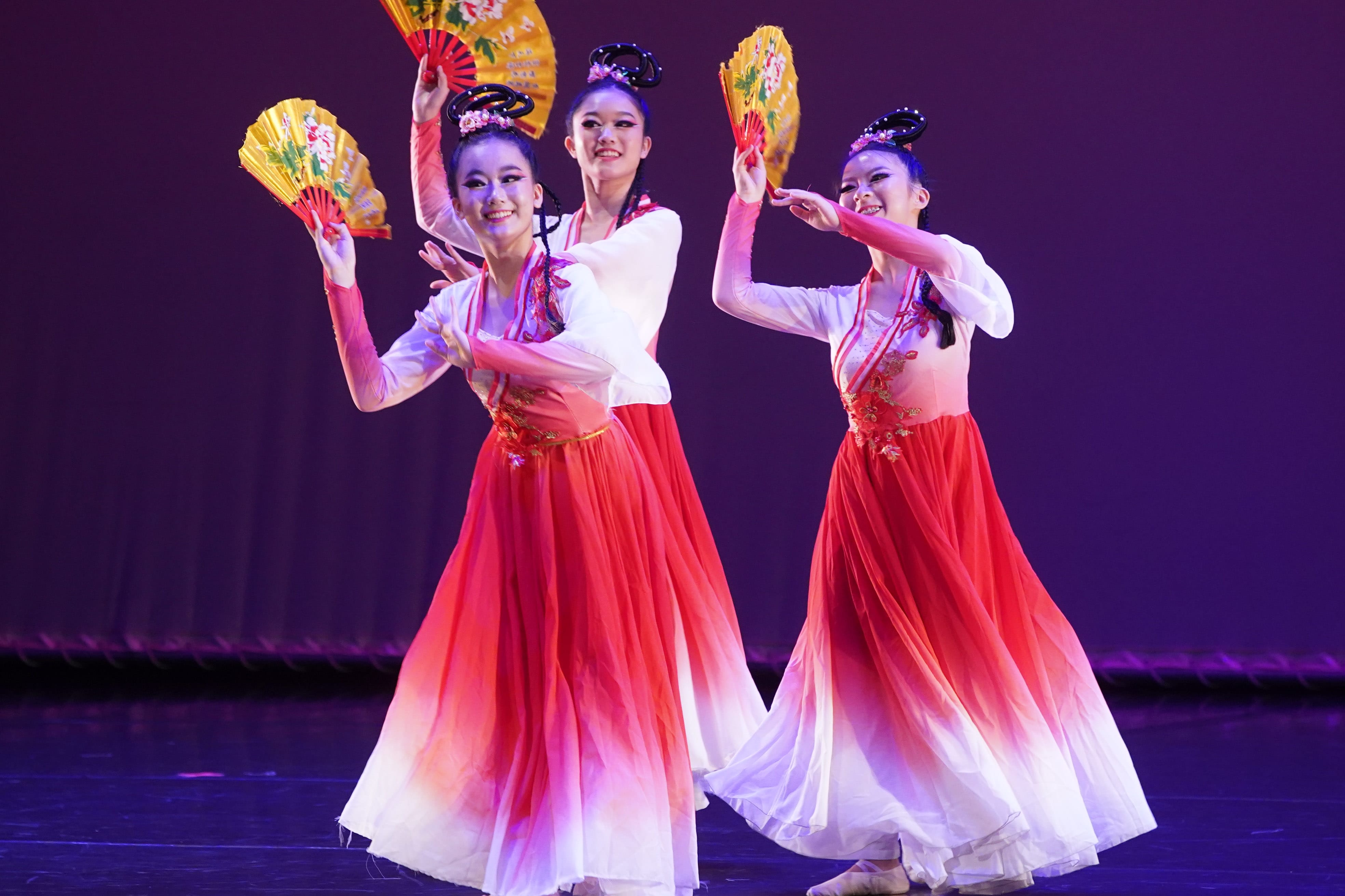 Dancers in traditional Chinese costumes perform a fan dance on a purple-lit stage.