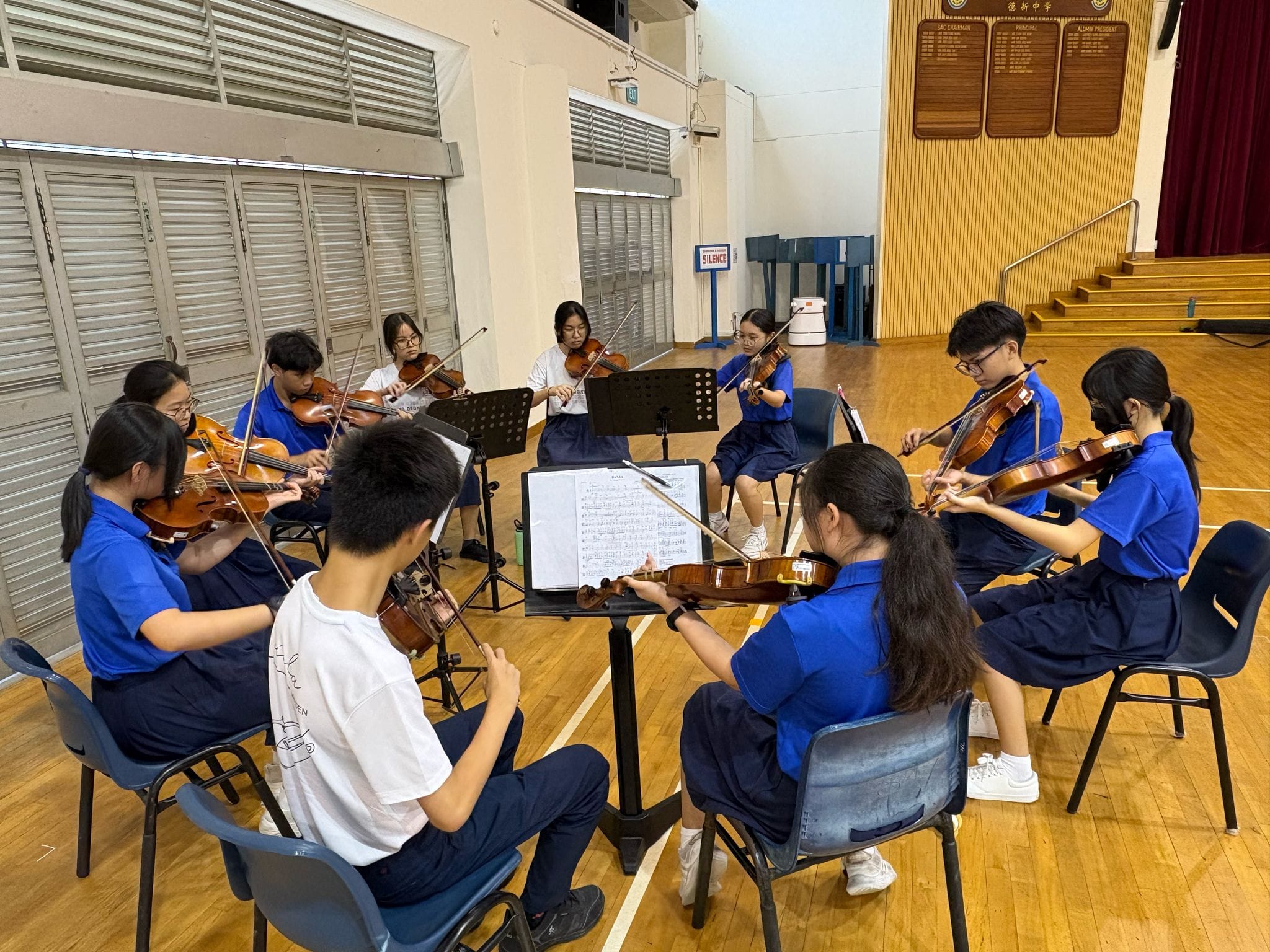 NV students sit in a circle playing violins in a school hall with music stands.
