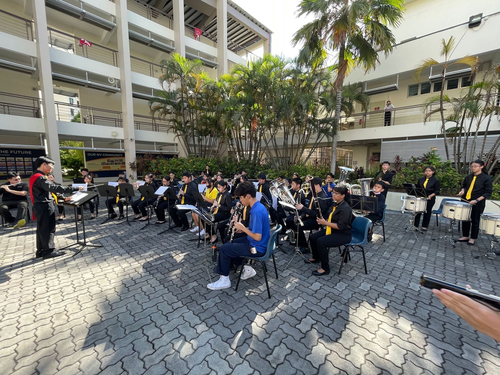 Band members perform outdoors in a school courtyard with a conductor leading them during National Day Celebration