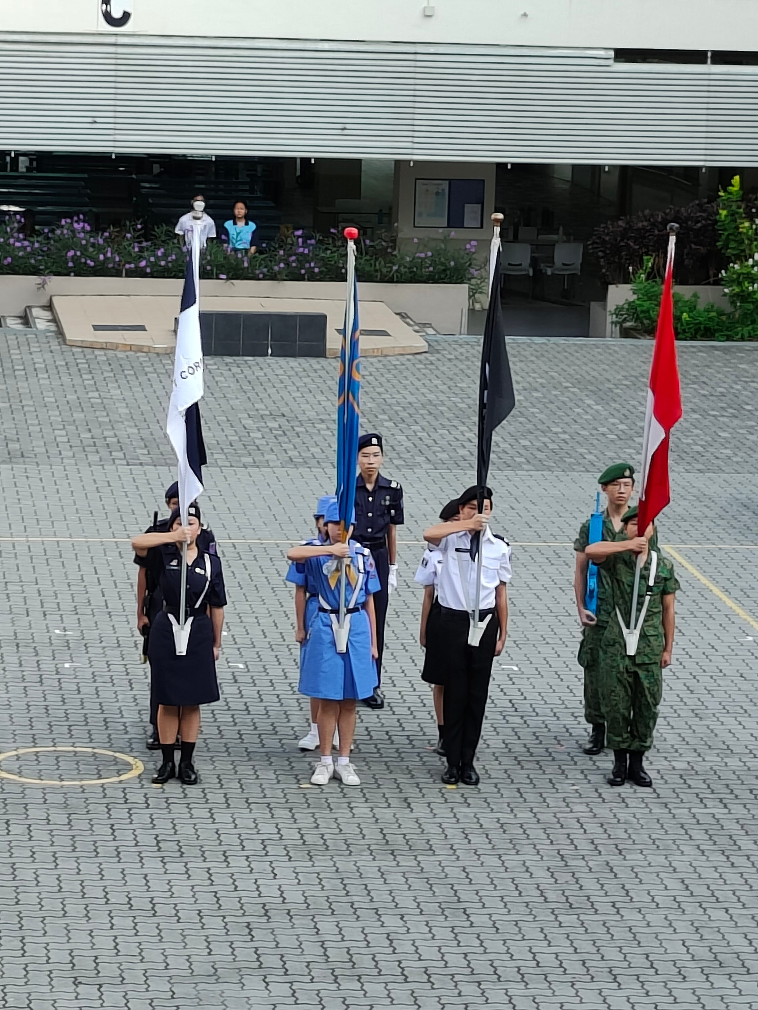 Students in various uniforms stand at attention on the parade square, holding colorful flags including Singapore flag.