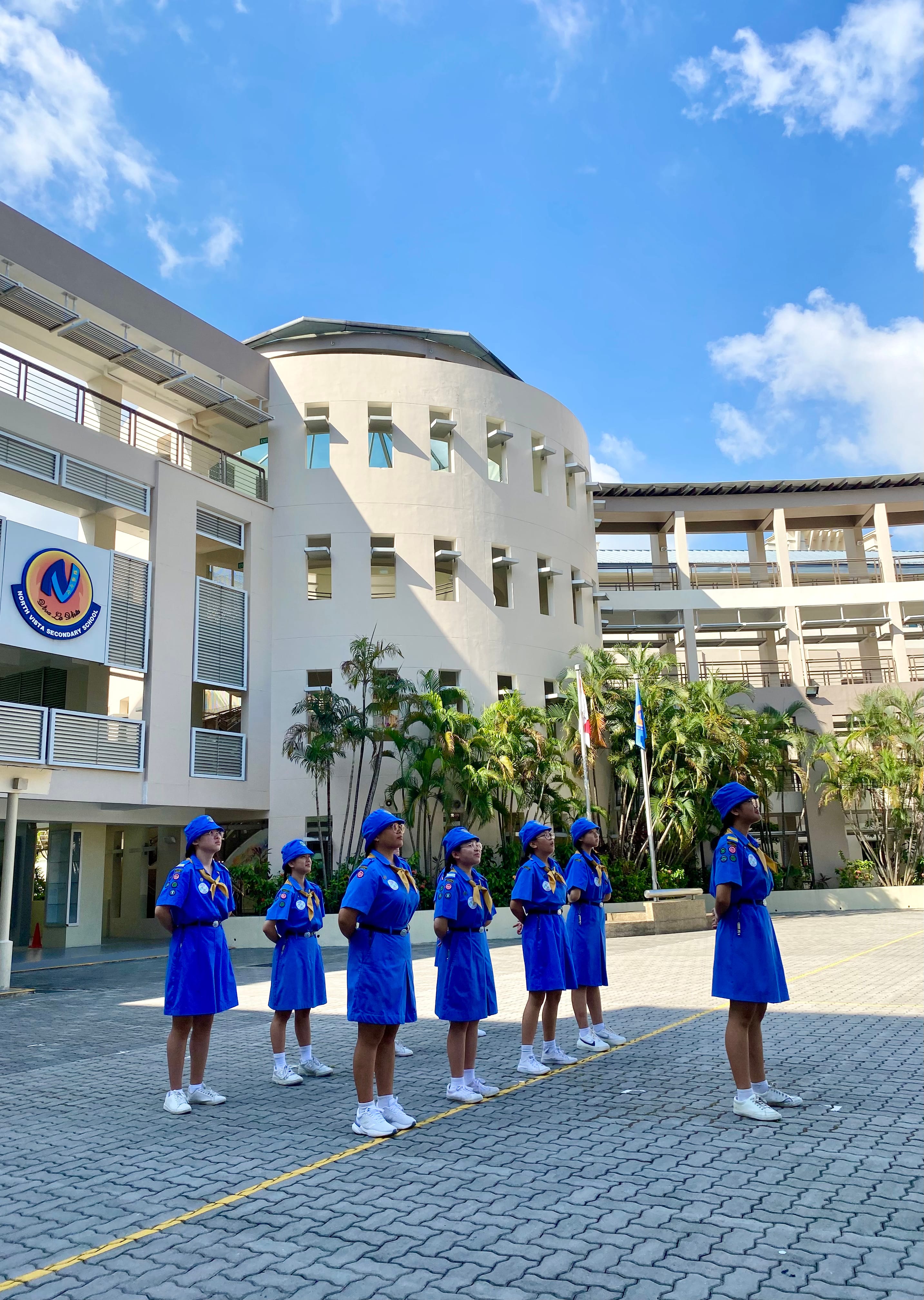 NV Girl Guides members stand at attention in a school parade square during a ceremony.
