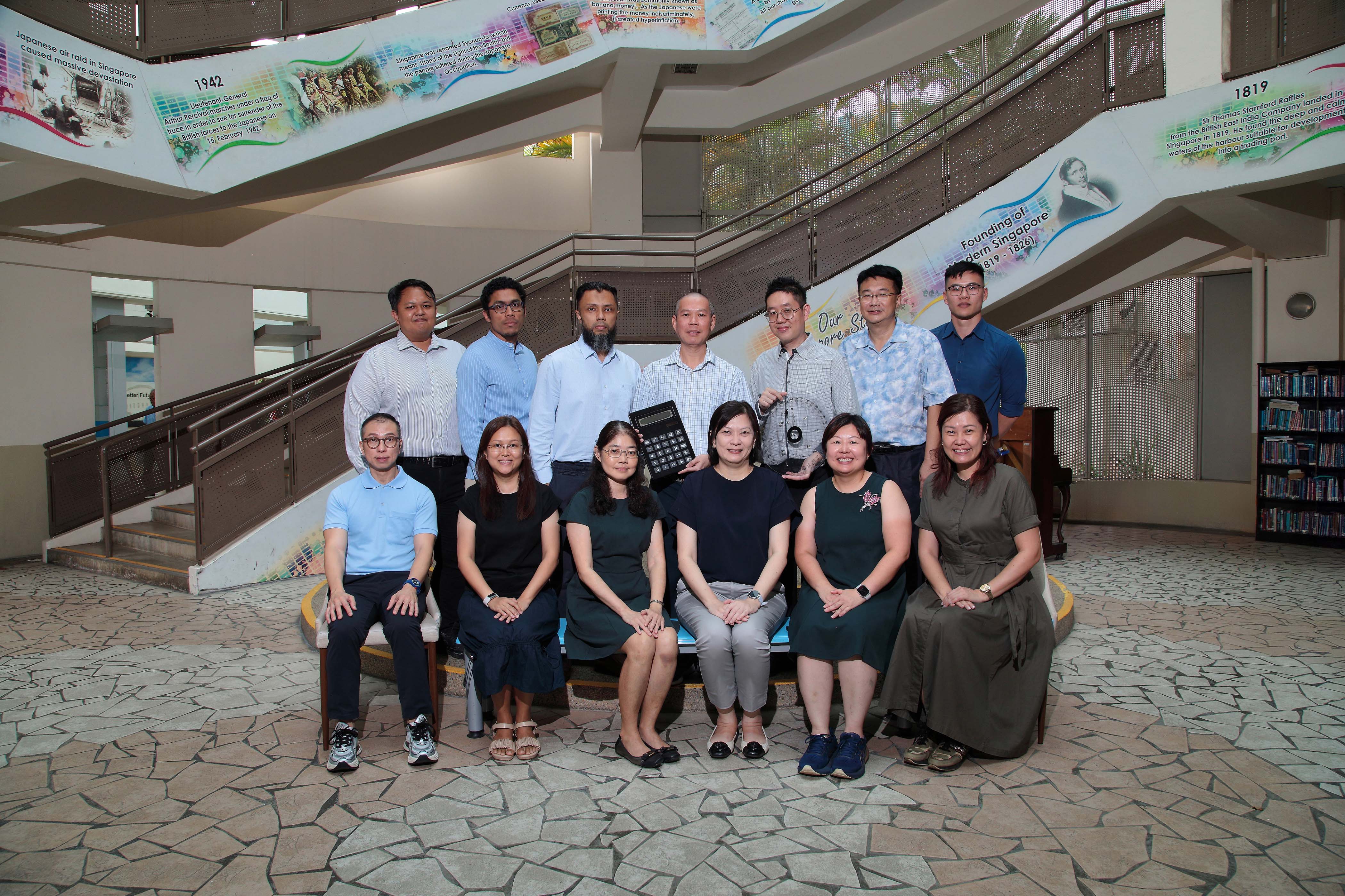 NV Math department's staff pose in two rows inside a building with historical murals and staircases; one holds a large calculator.
