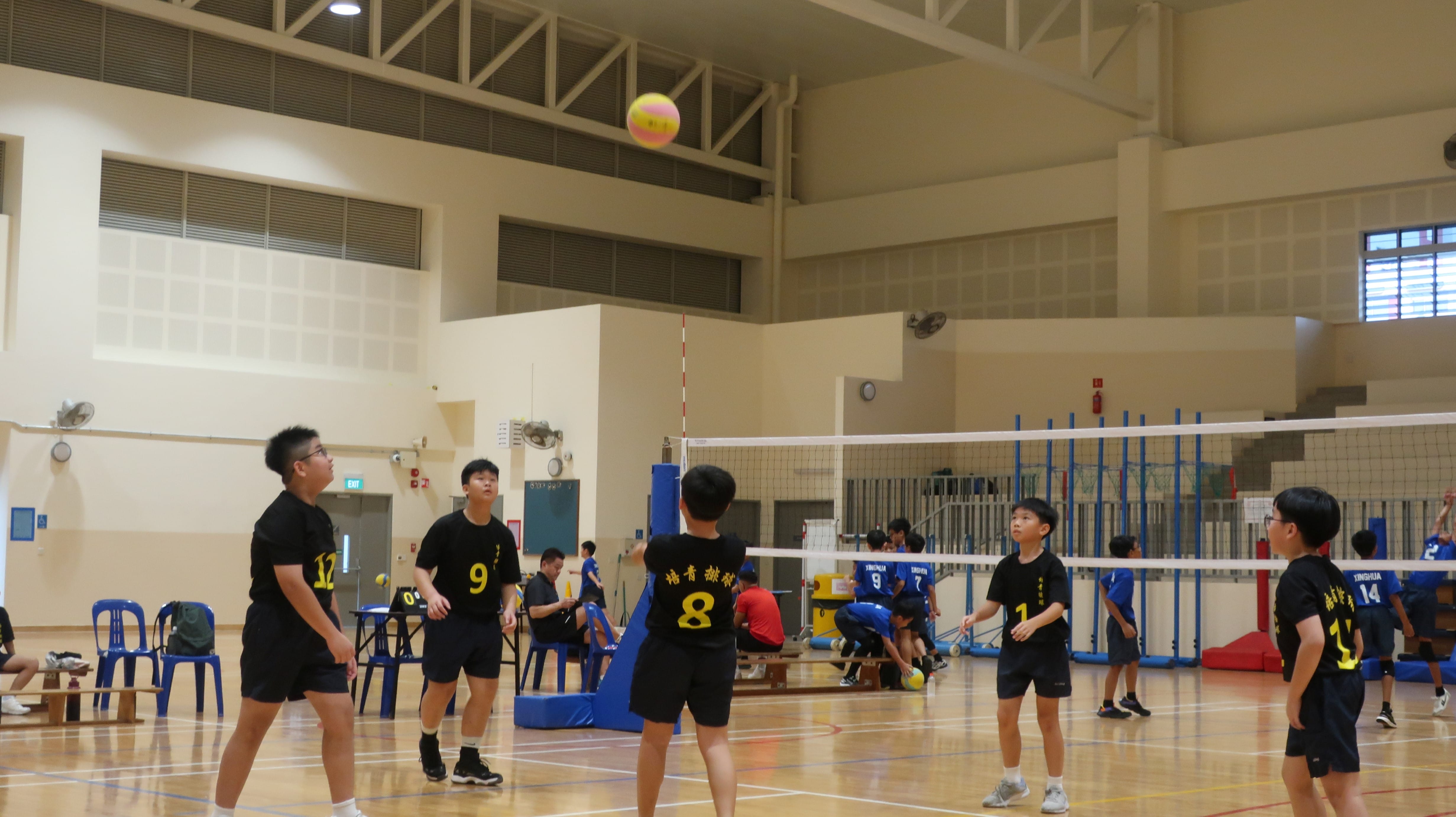 Young players in black jerseys play volleyball indoors, with a yellow-pink ball mid-air above the net.