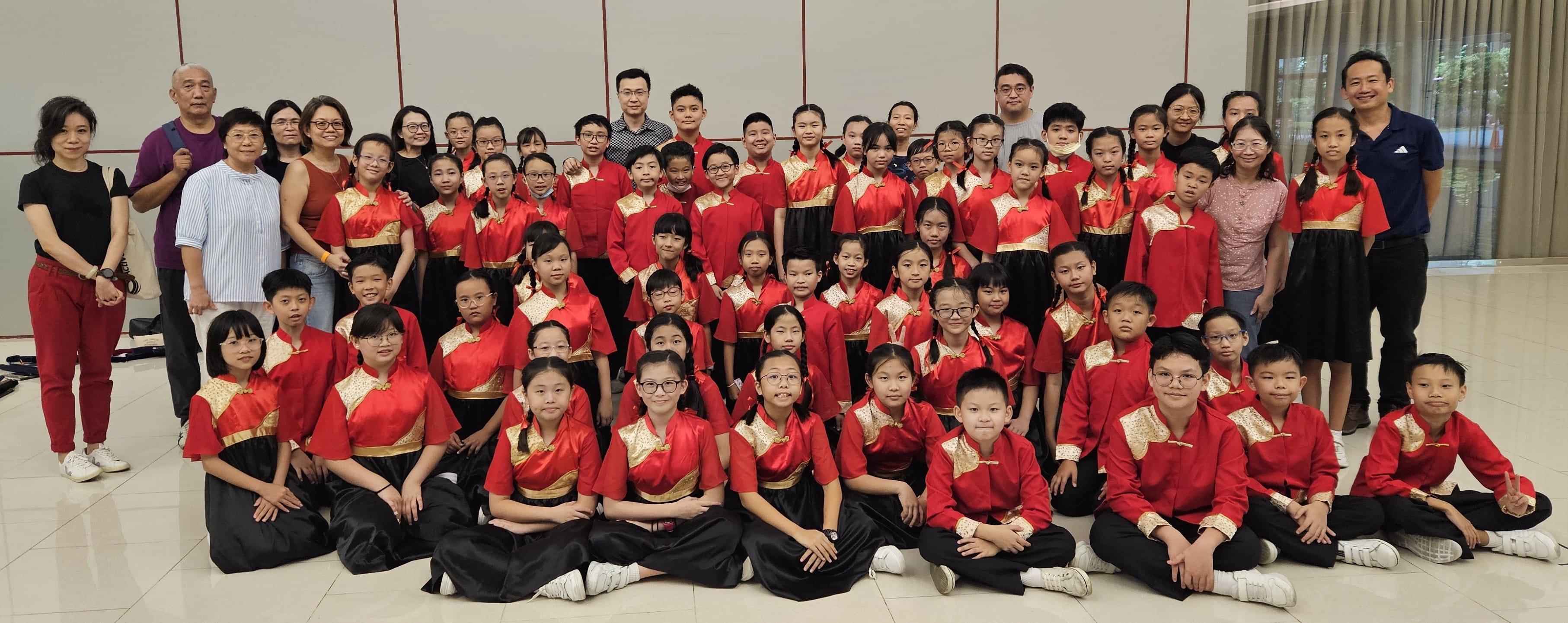 Large group of children in red and gold traditional Chinese costumes posing with adults for a group photo indoors.