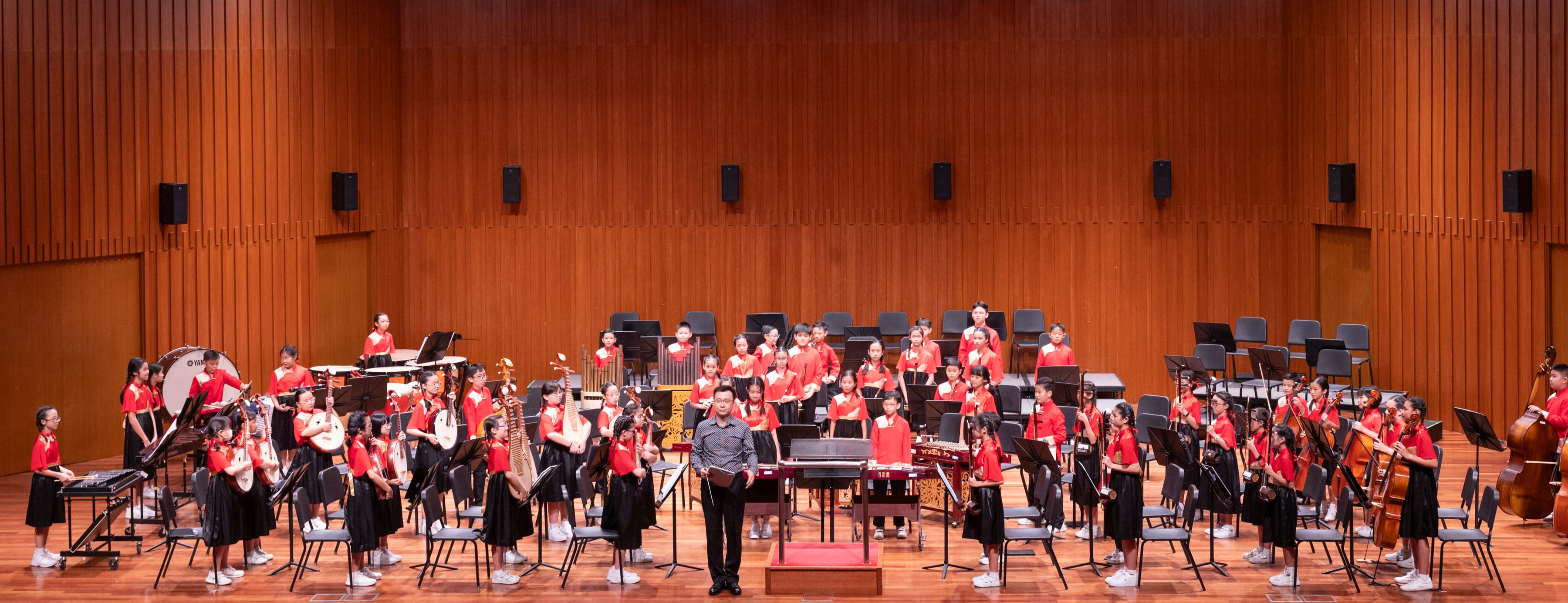 A conductor stands before a large youth orchestra of children in red uniforms on a wooden concert hall stage.