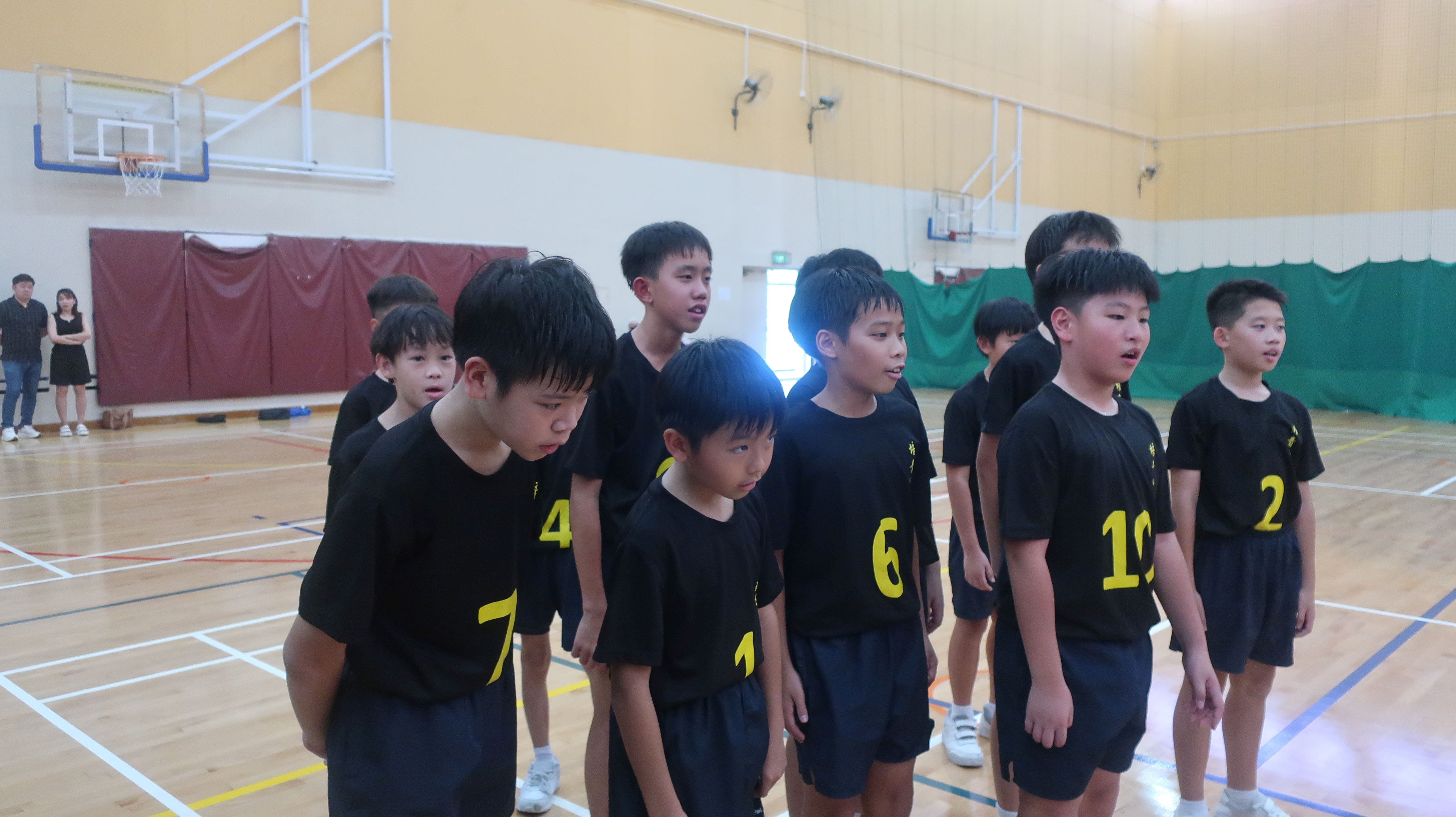 A group of young boys in black jerseys with yellow numbers stand in a gymnasium with basketball hoops.