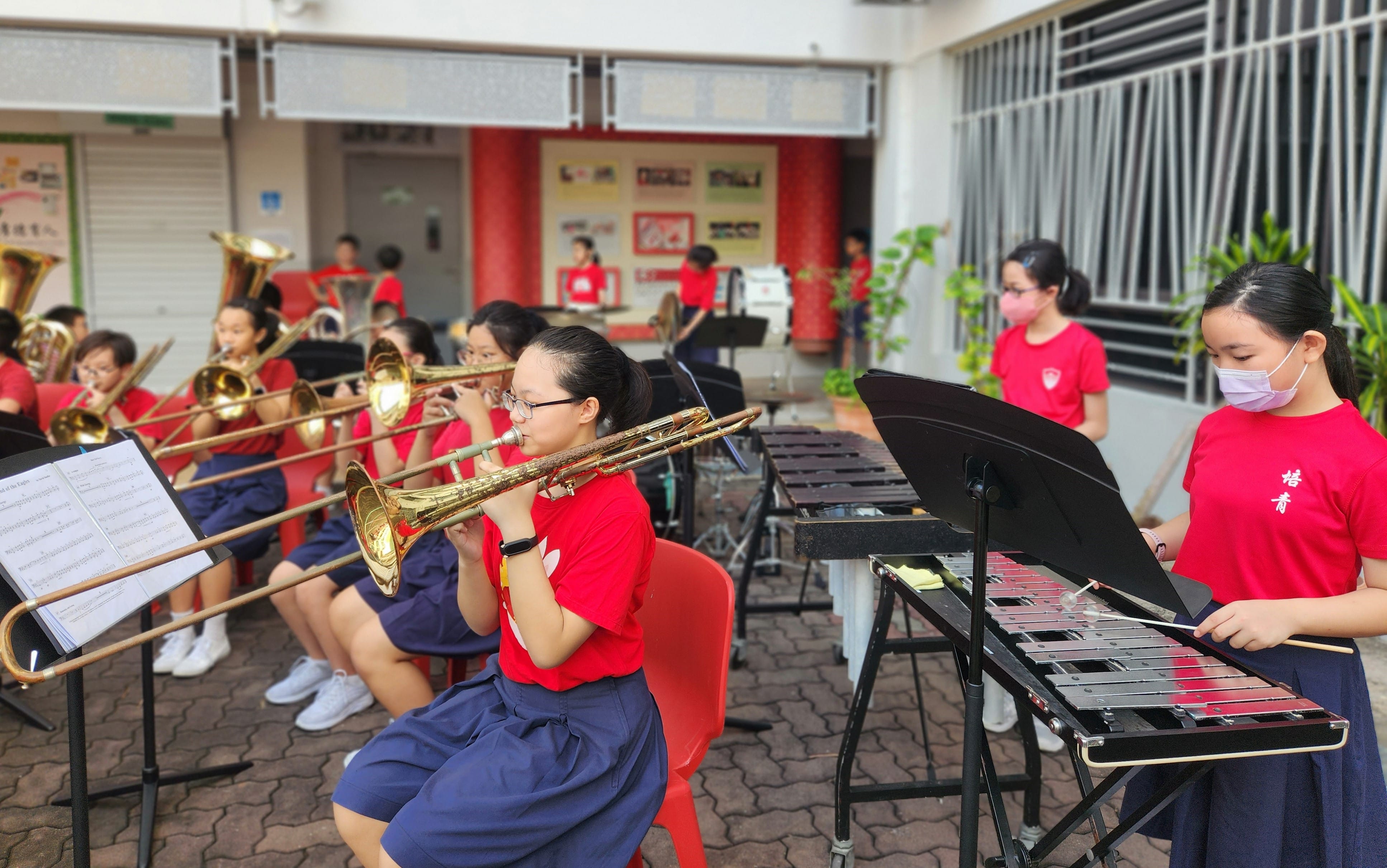 Students in red shirts play brass instruments and a xylophone outdoors during a school band rehearsal.