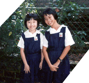 Two young girls wearing matching navy blue pinafore uniforms with white shirts, smiling outdoors near green foliage.