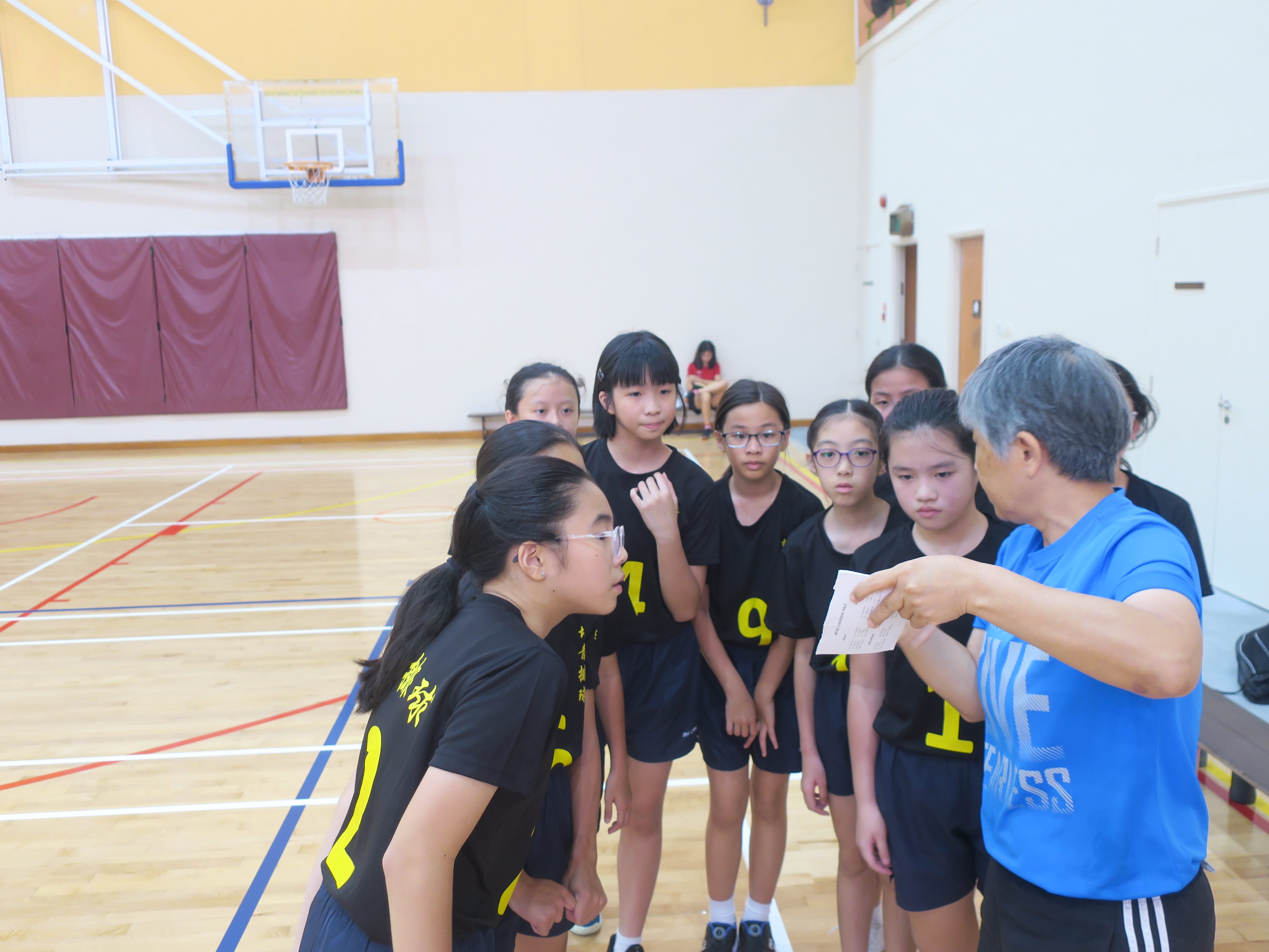 A coach in a blue shirt holds a paper and talks to a group of young girls in black jerseys on a basketball court.