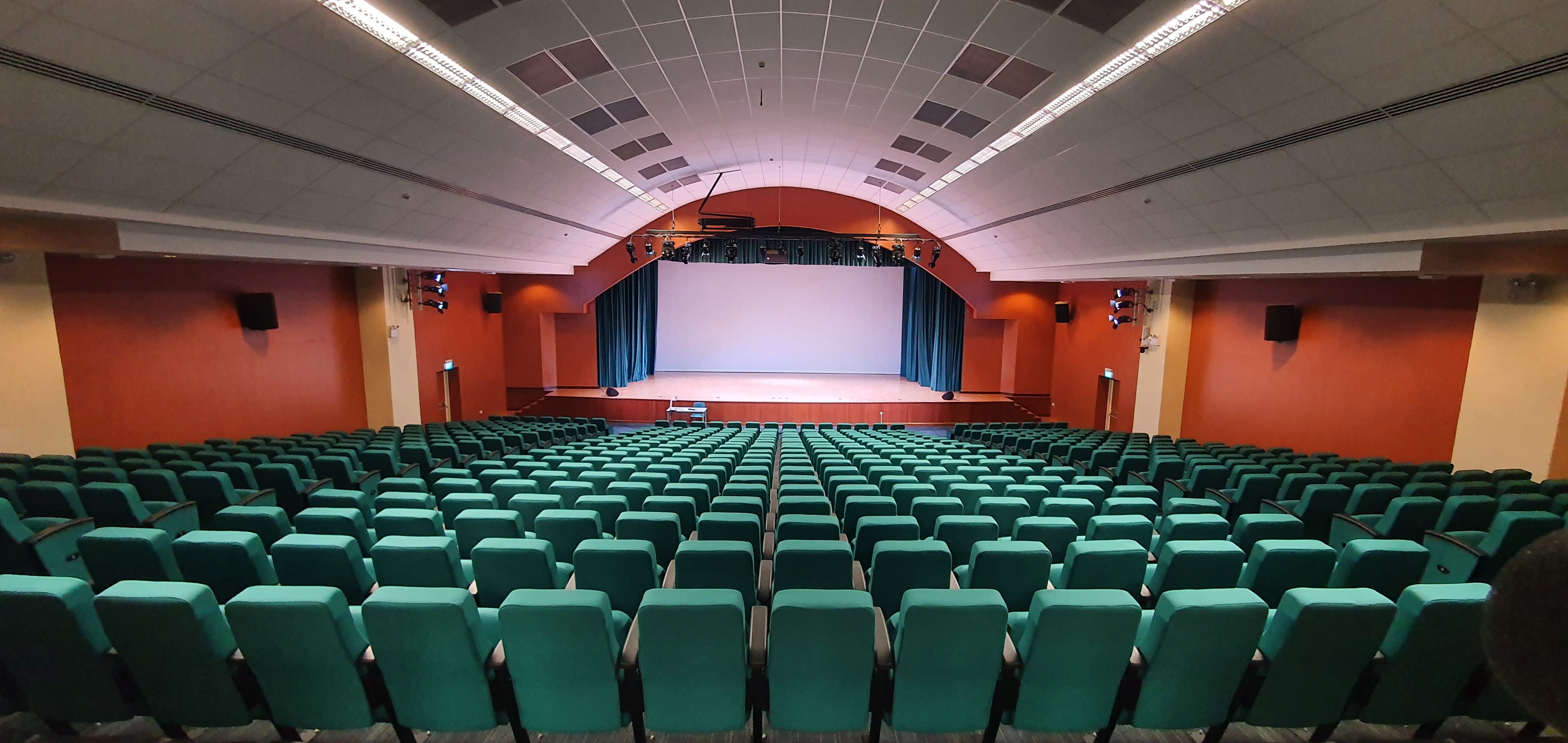 Empty auditorium with rows of green seats facing a stage with blue curtains and a white screen.