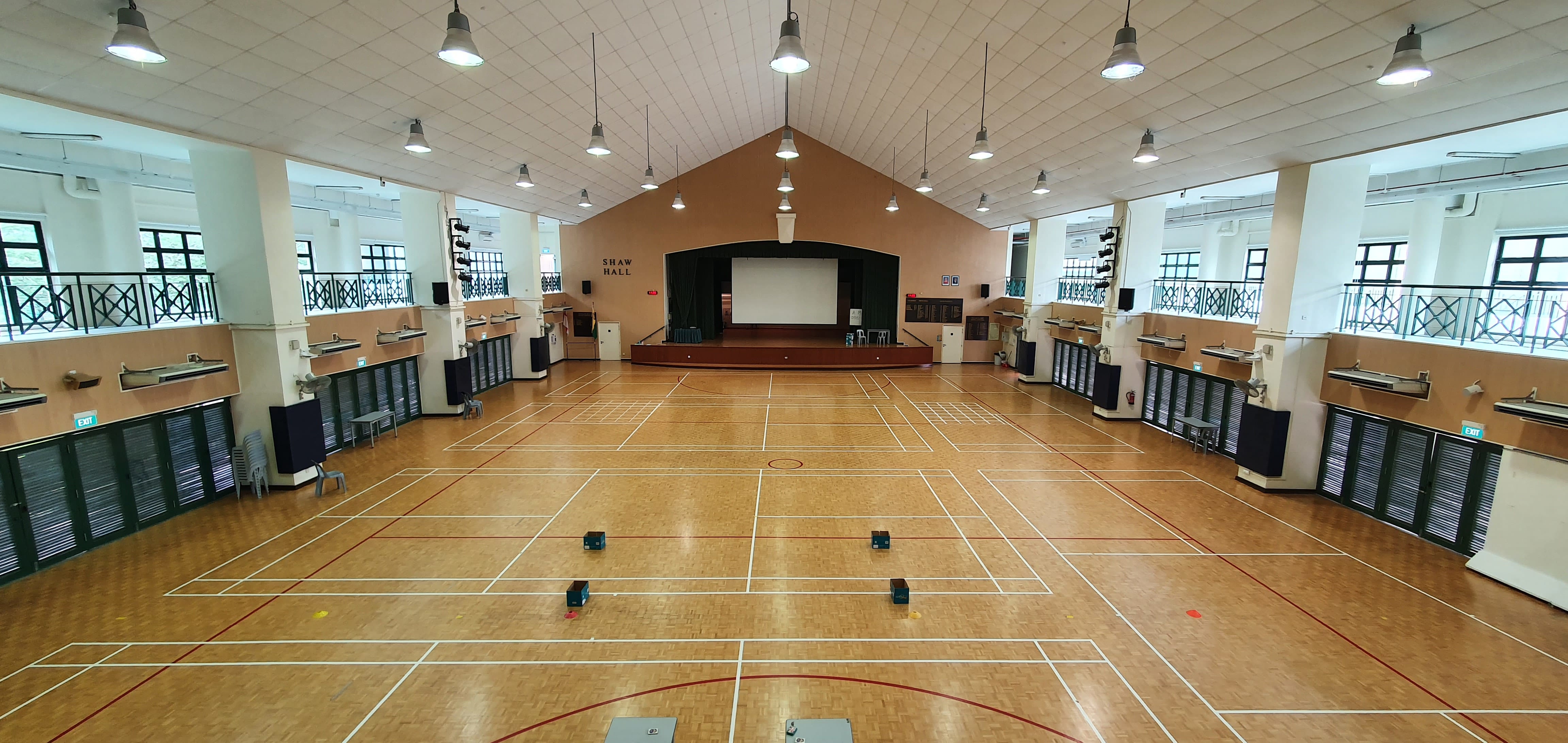 Large empty indoor hall named Shaw Hall with a wooden court floor, raised stage, projection screen, and pendant lights.