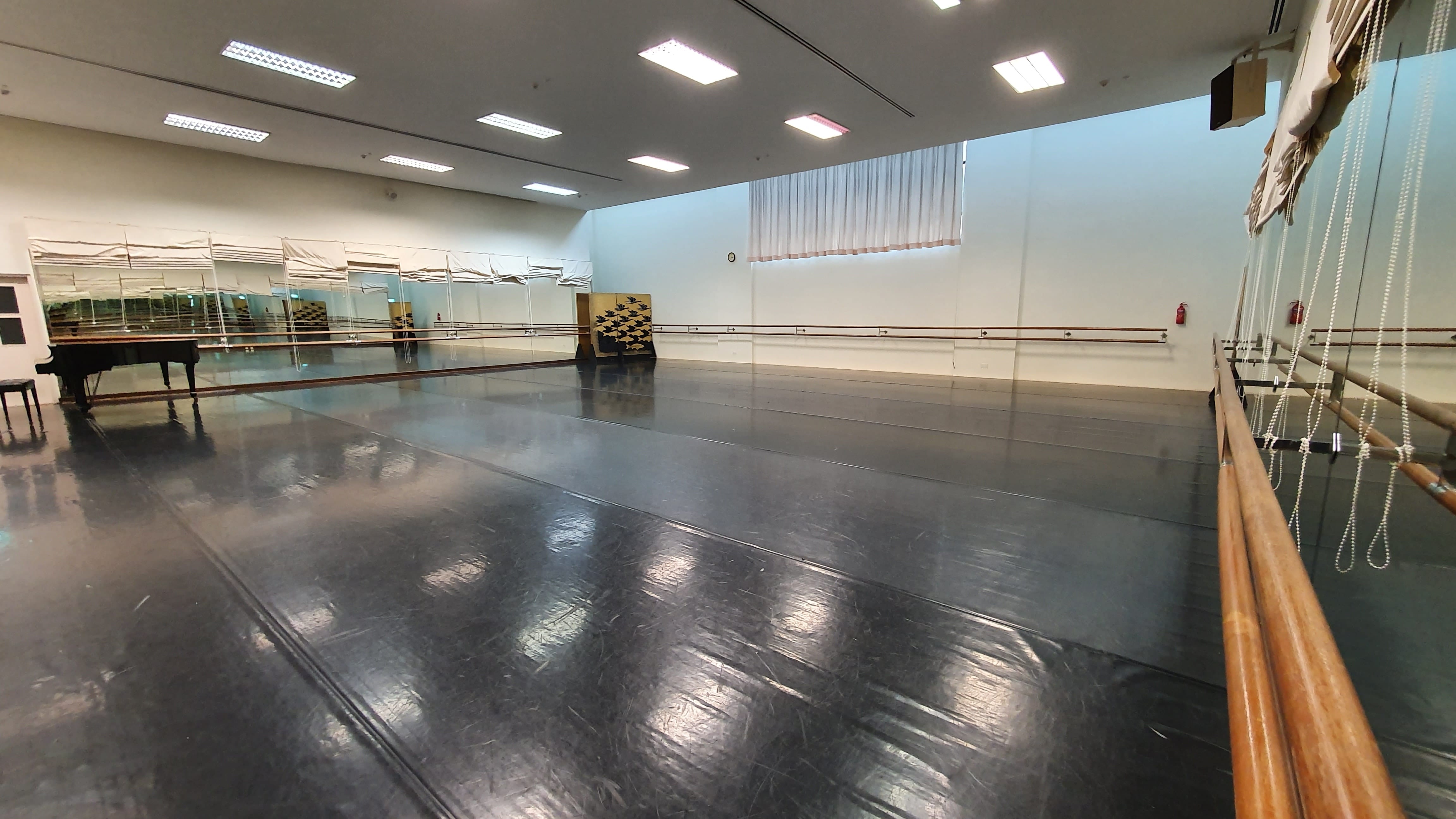 Empty ballet studio with dark floor, wooden barres on walls, large mirrors, and a grand piano in the corner.