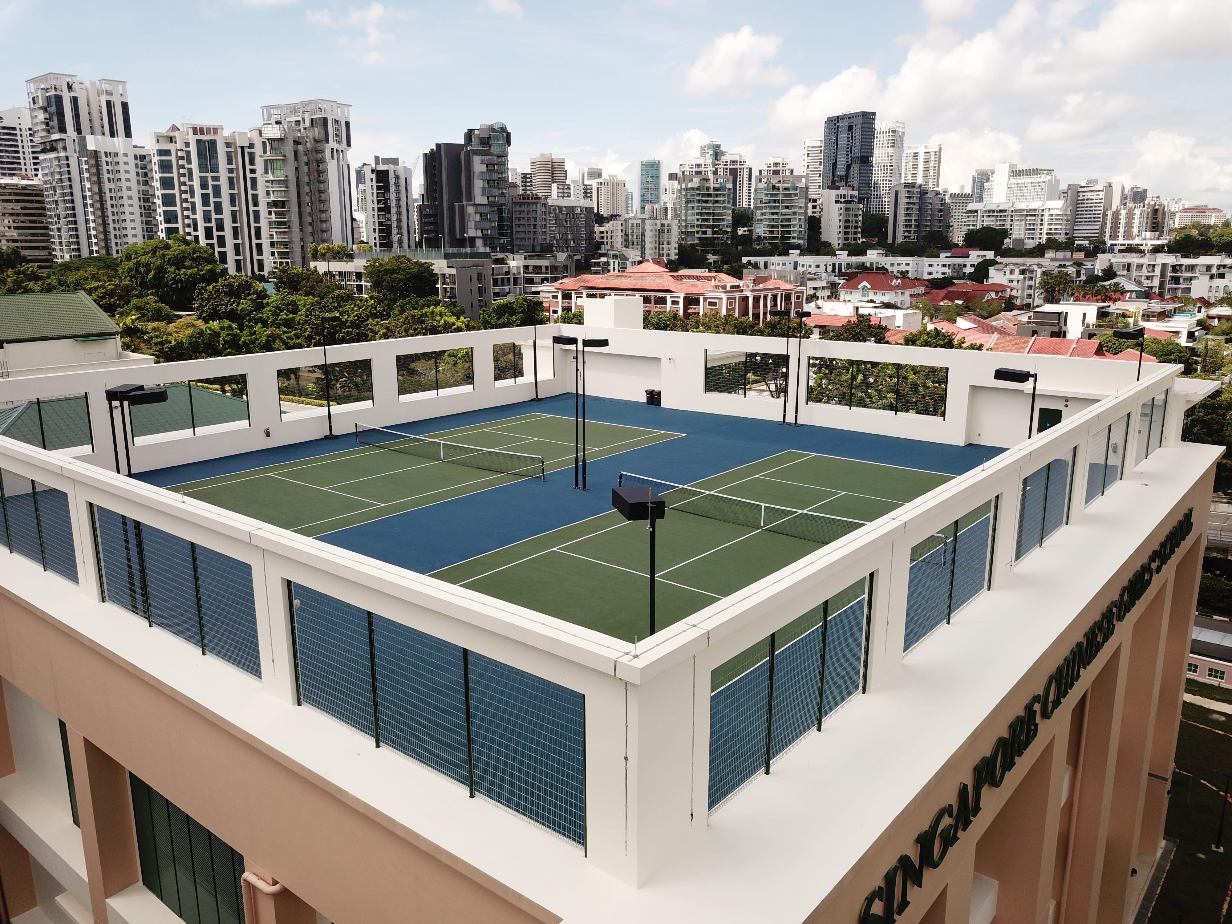Rooftop tennis courts with nets and lights on a building, city skyline and trees in the background.