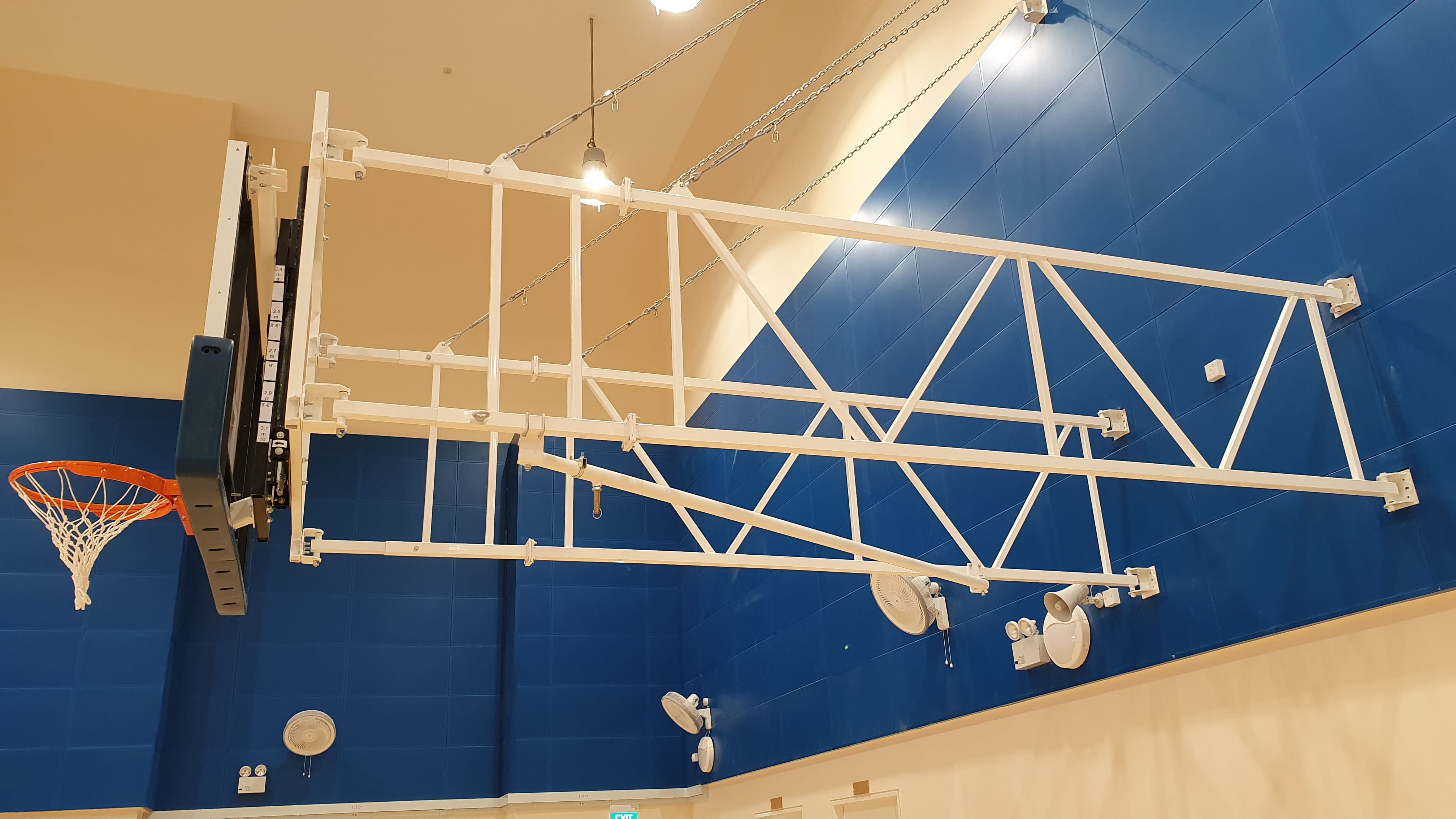 A wall-mounted basketball hoop with white metal truss arm extends along a blue tiled indoor gym wall.