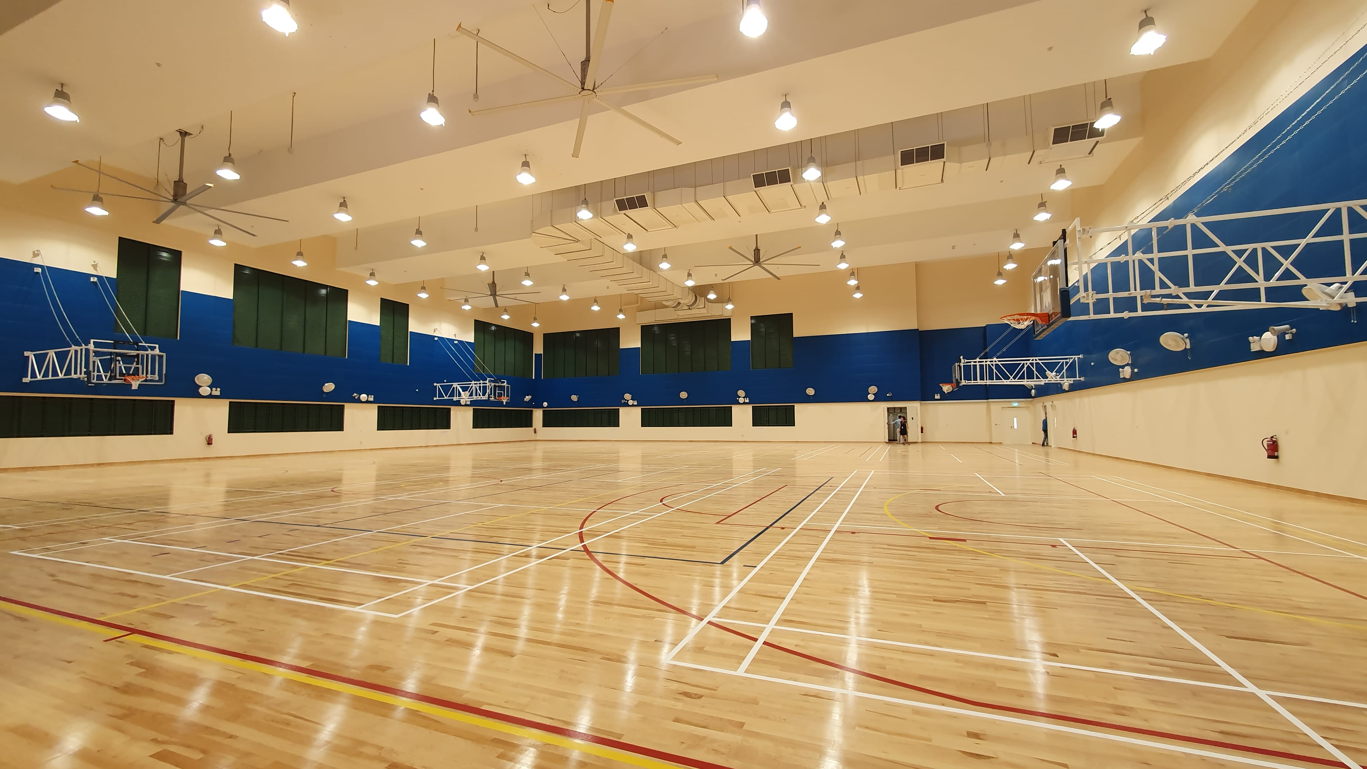 Large indoor sports hall with a polished wood floor, colorful court lines, basketball hoops, blue walls, and ceiling fans.