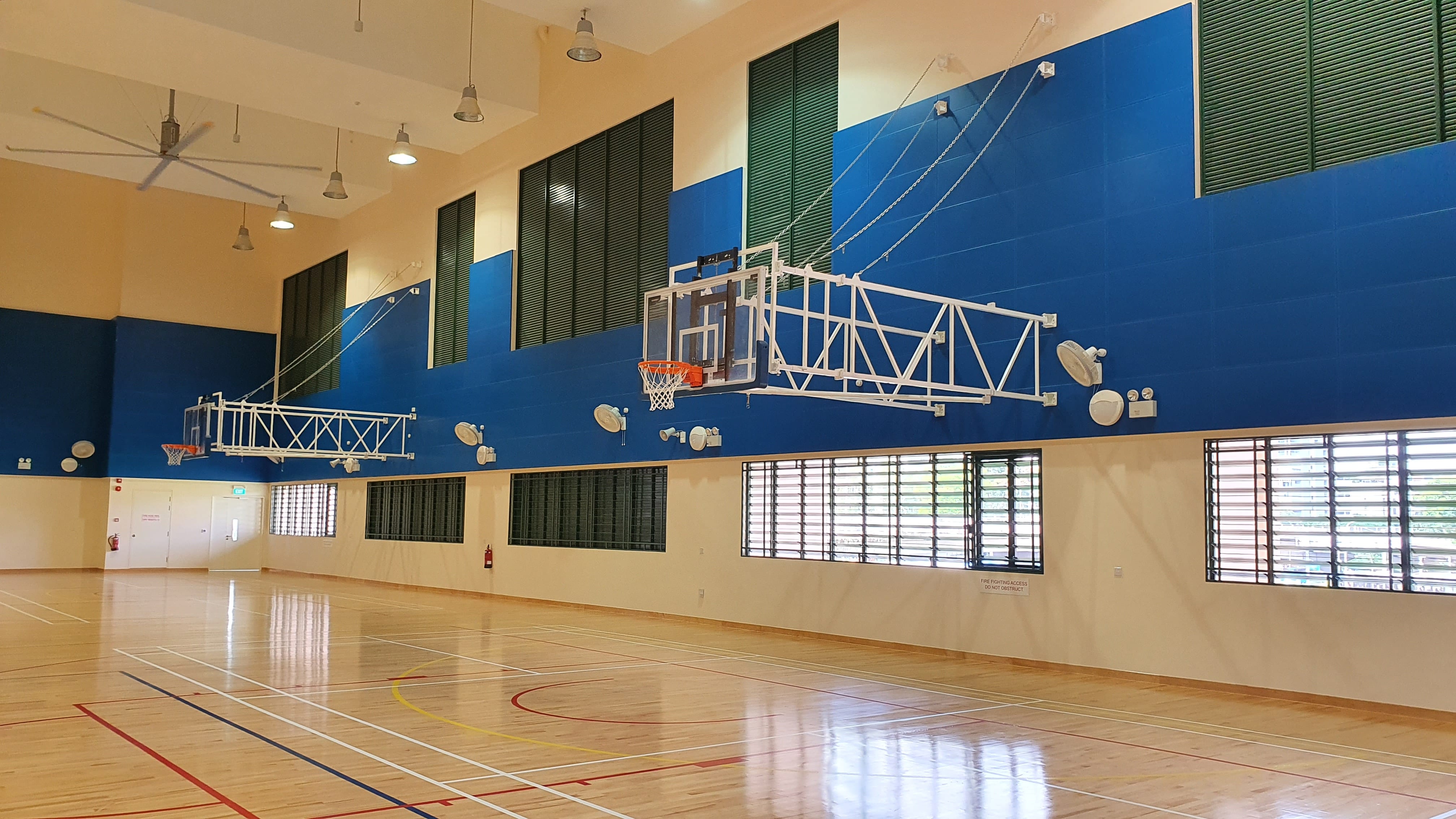 Empty indoor basketball court with polished wood floor, blue walls, wall-mounted hoops, and ceiling fans.