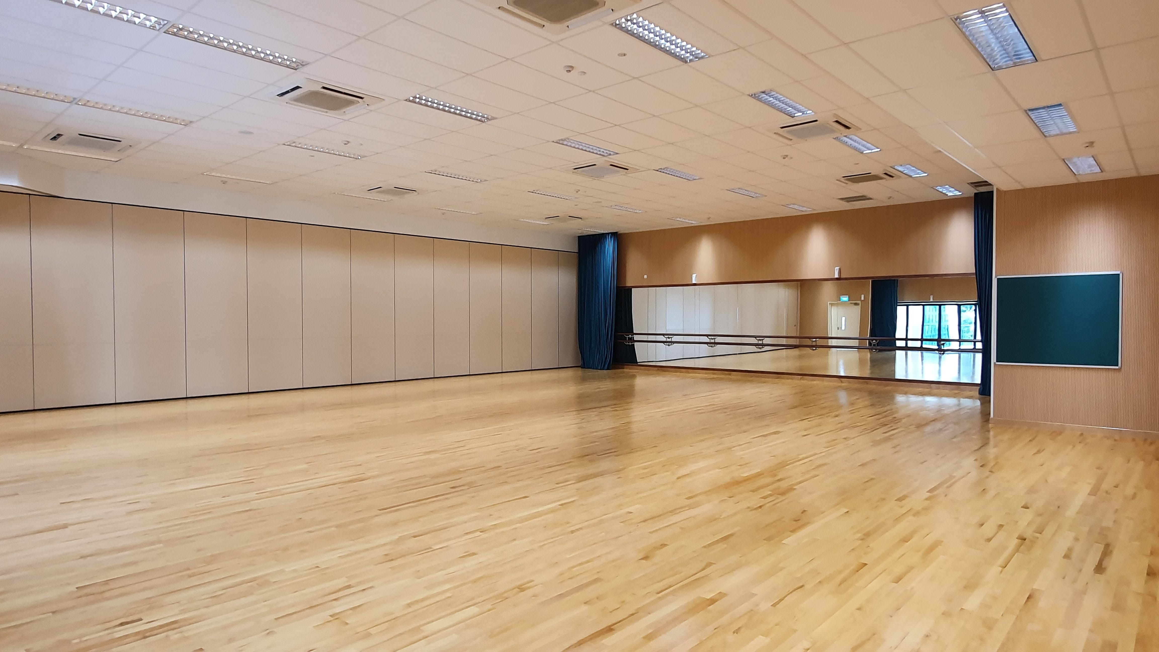 Empty dance studio with wooden floor, wall mirrors, ballet barres, folding partition walls, and fluorescent ceiling lights.