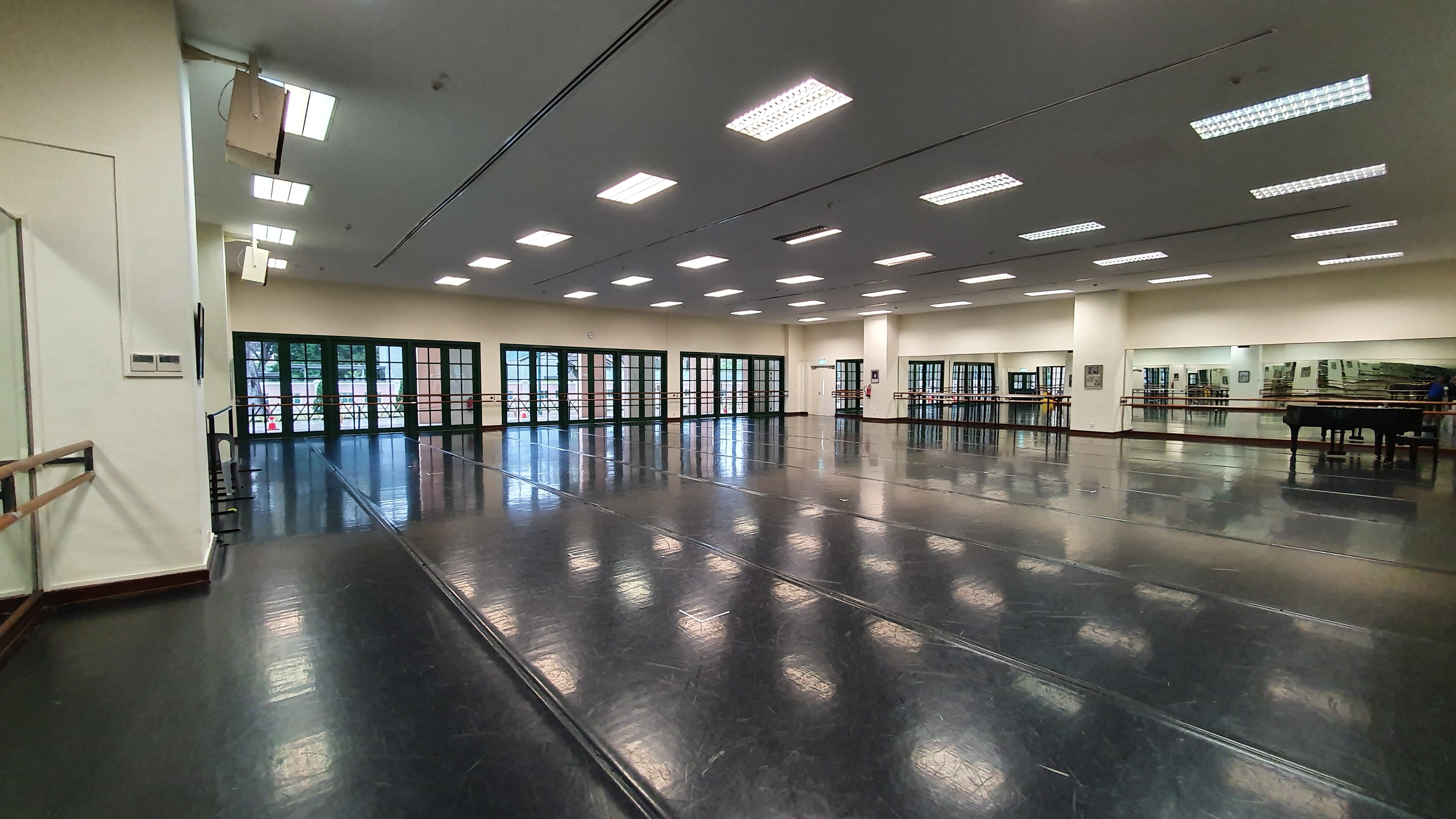 Large empty dance studio with dark polished floor, wall mirrors, ballet barres, a grand piano, and glass-paneled doors.