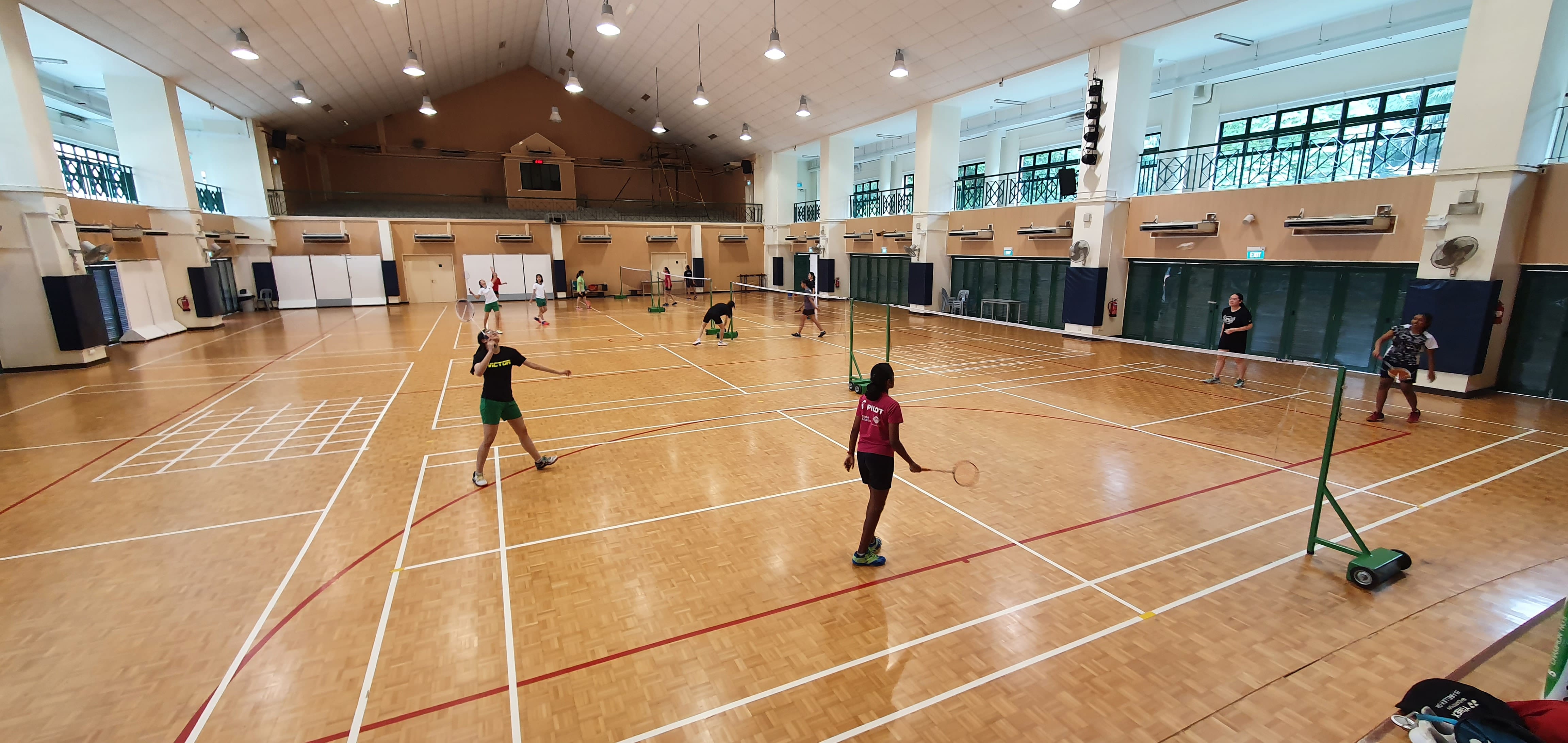 Several players practice badminton on multiple courts inside a large indoor sports hall with wooden flooring.