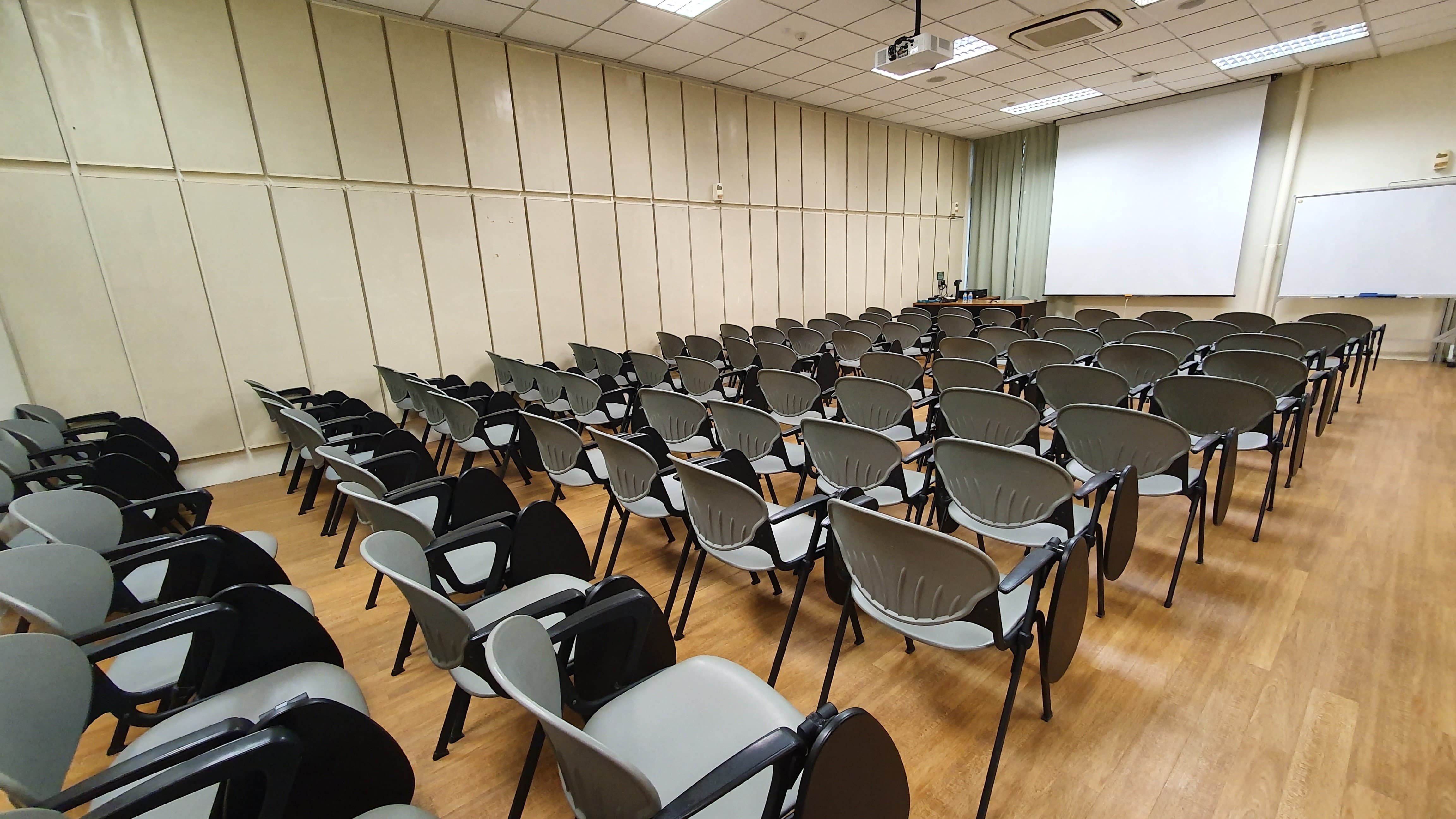 Empty lecture room with rows of gray chairs, a projector on the ceiling, and a white screen at the front.