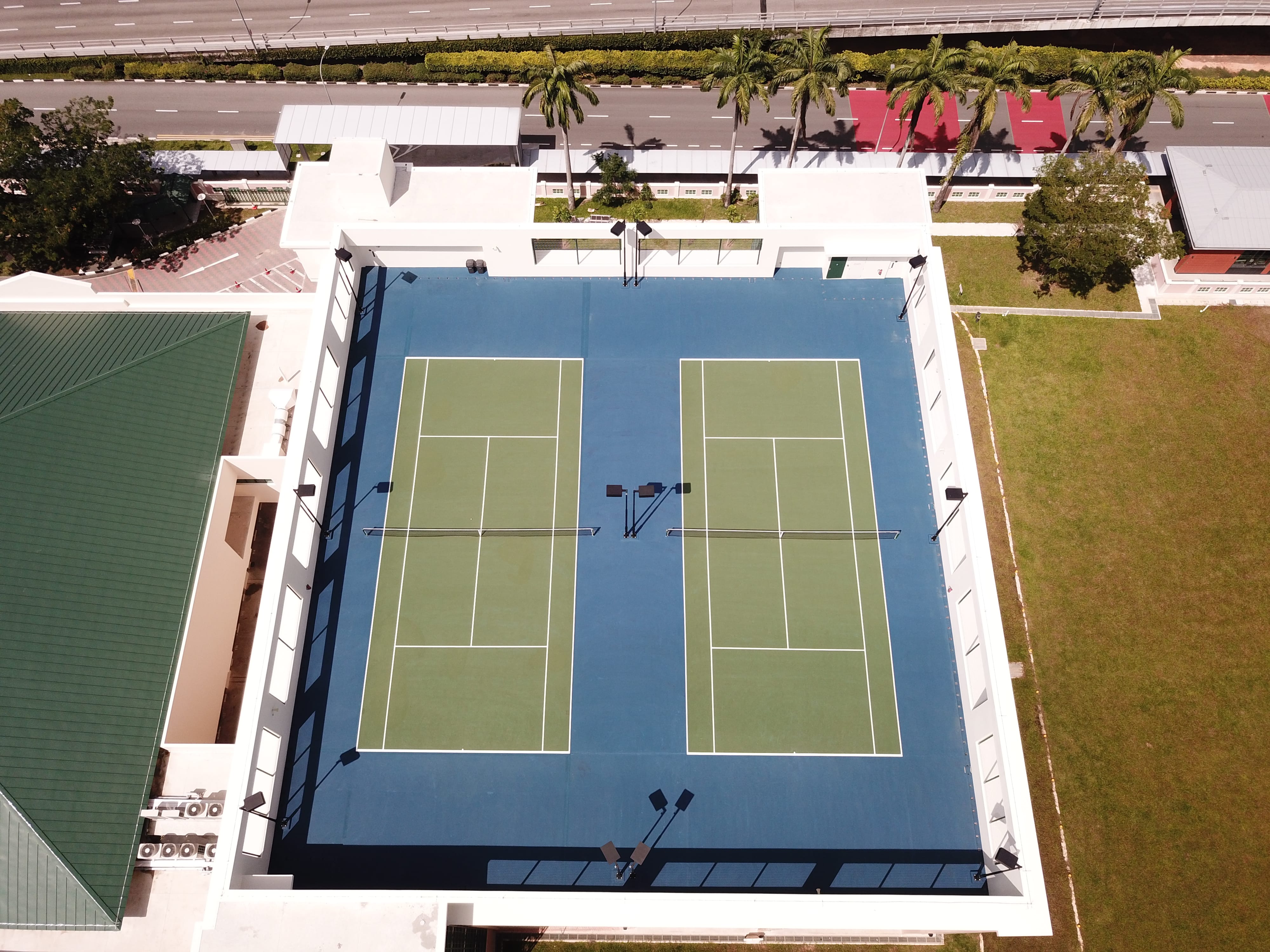 Aerial view of two adjacent tennis courts in a walled complex, with palm trees and a road above.