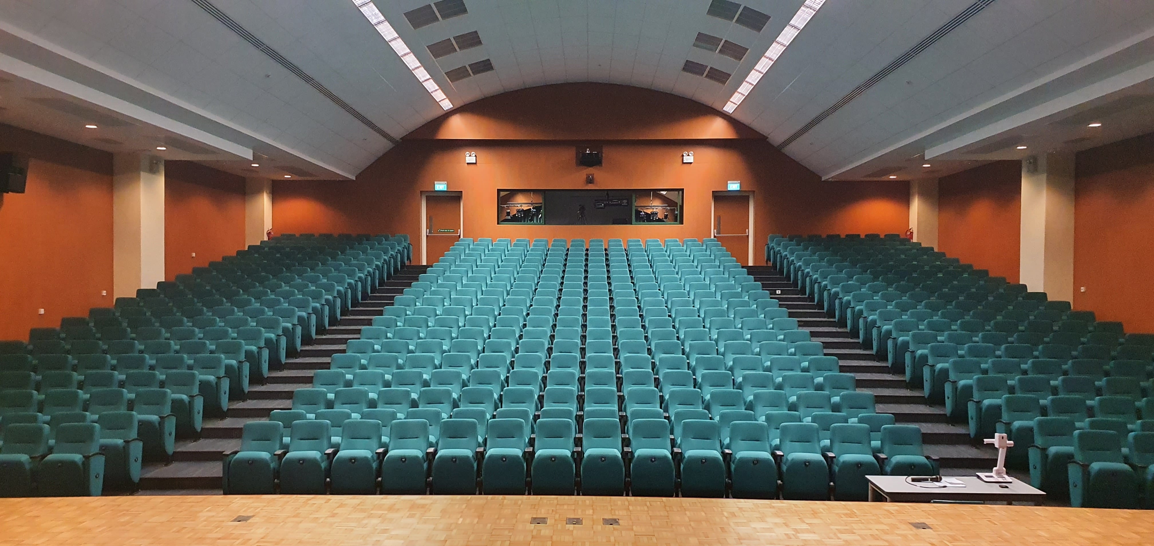 Empty auditorium with rows of teal seats, orange walls, arched ceiling, and a wooden stage in the foreground.