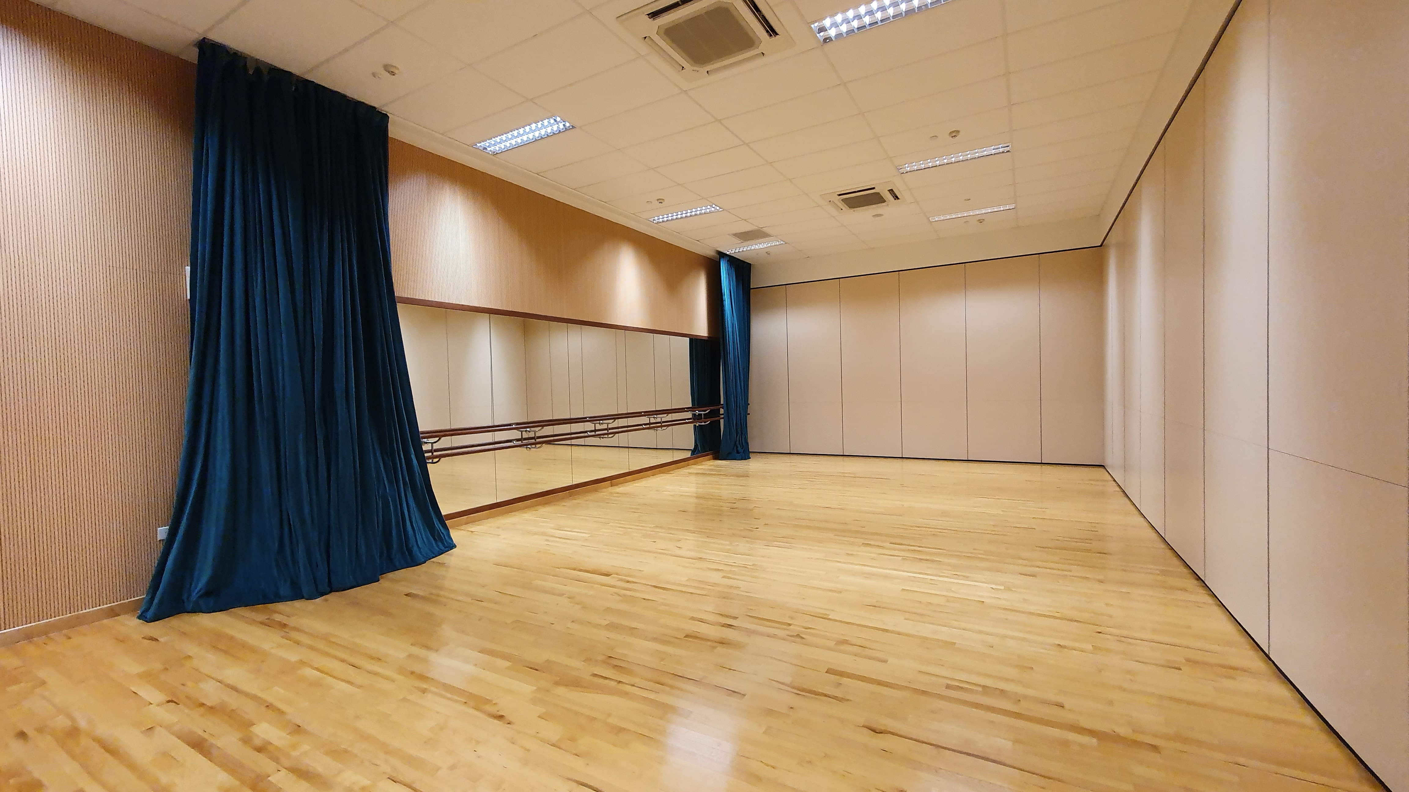 Empty dance studio with wood floor, wall mirrors, ballet barres, blue curtains, and ceiling lights.