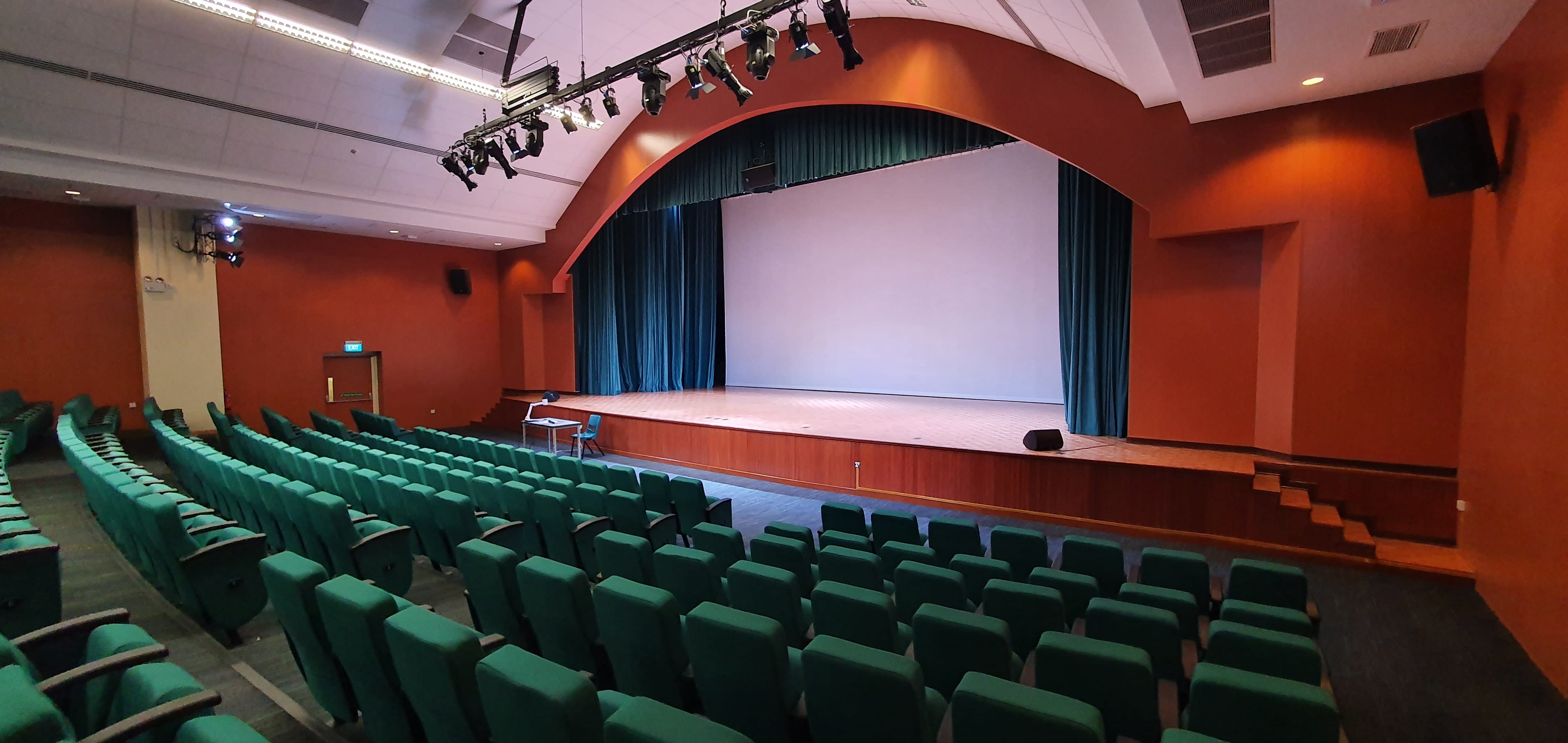Empty auditorium with green seats, a wooden stage, teal curtains, a white screen, and overhead stage lighting.