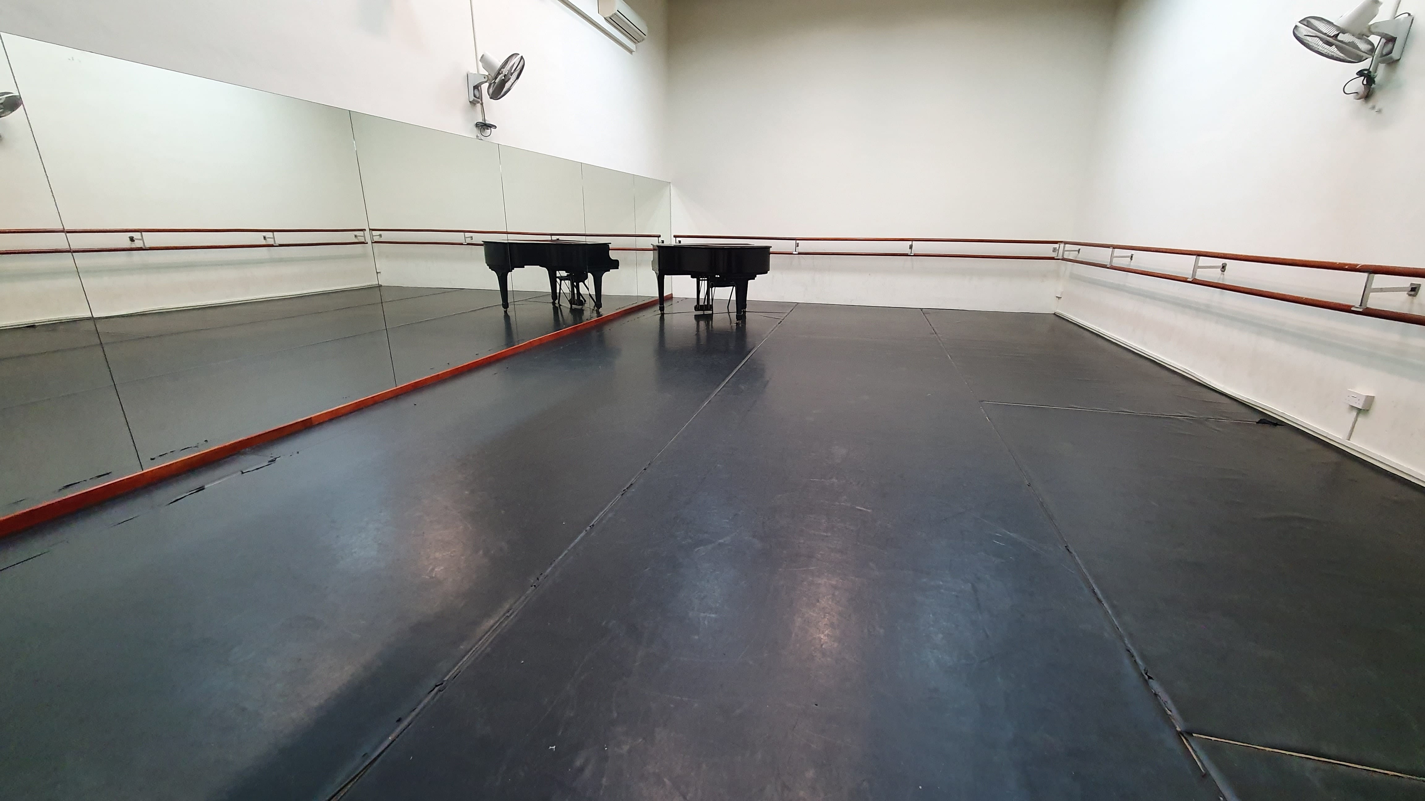 Empty dance studio with dark floor, wall mirrors, wooden ballet barre, a grand piano, and wall-mounted fans.