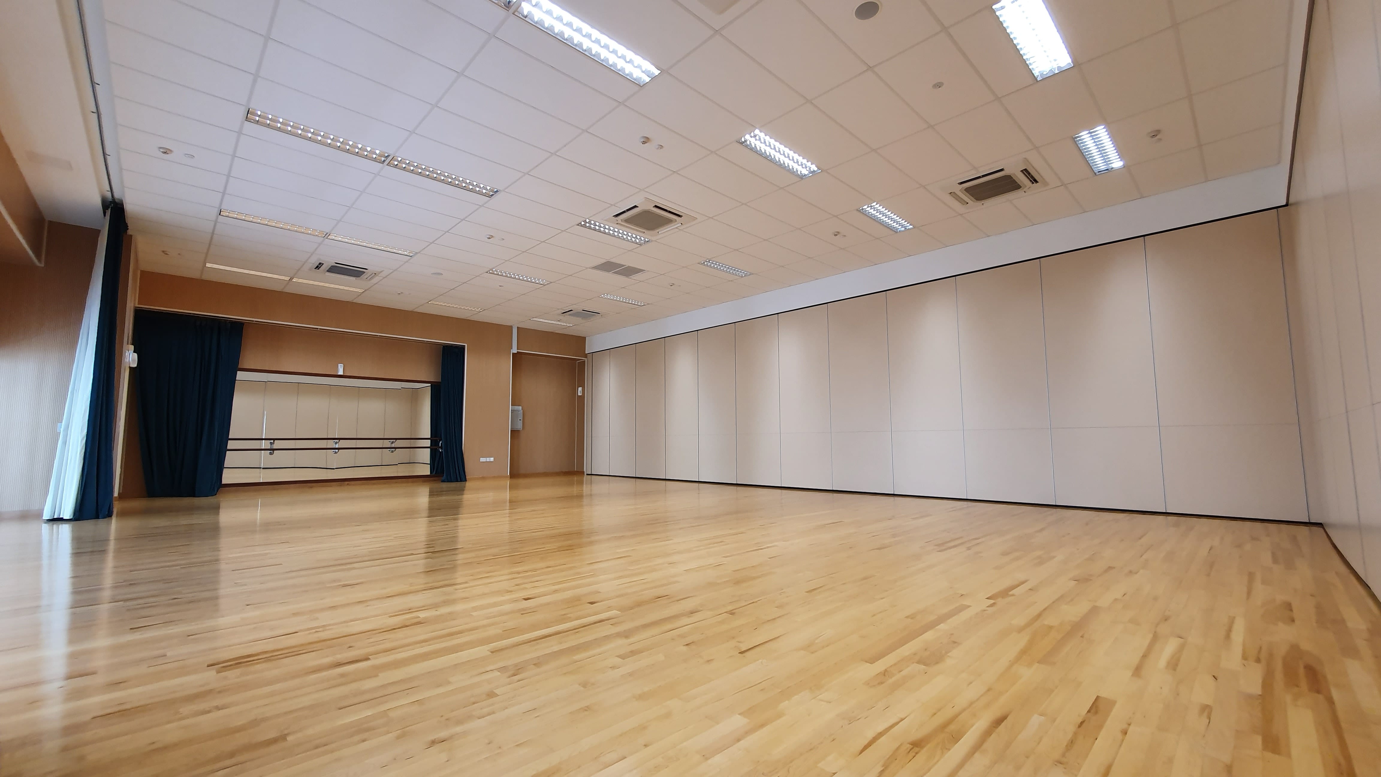Empty dance studio with polished wood floor, wall mirrors, ballet barres, blue curtains, and white partition walls.