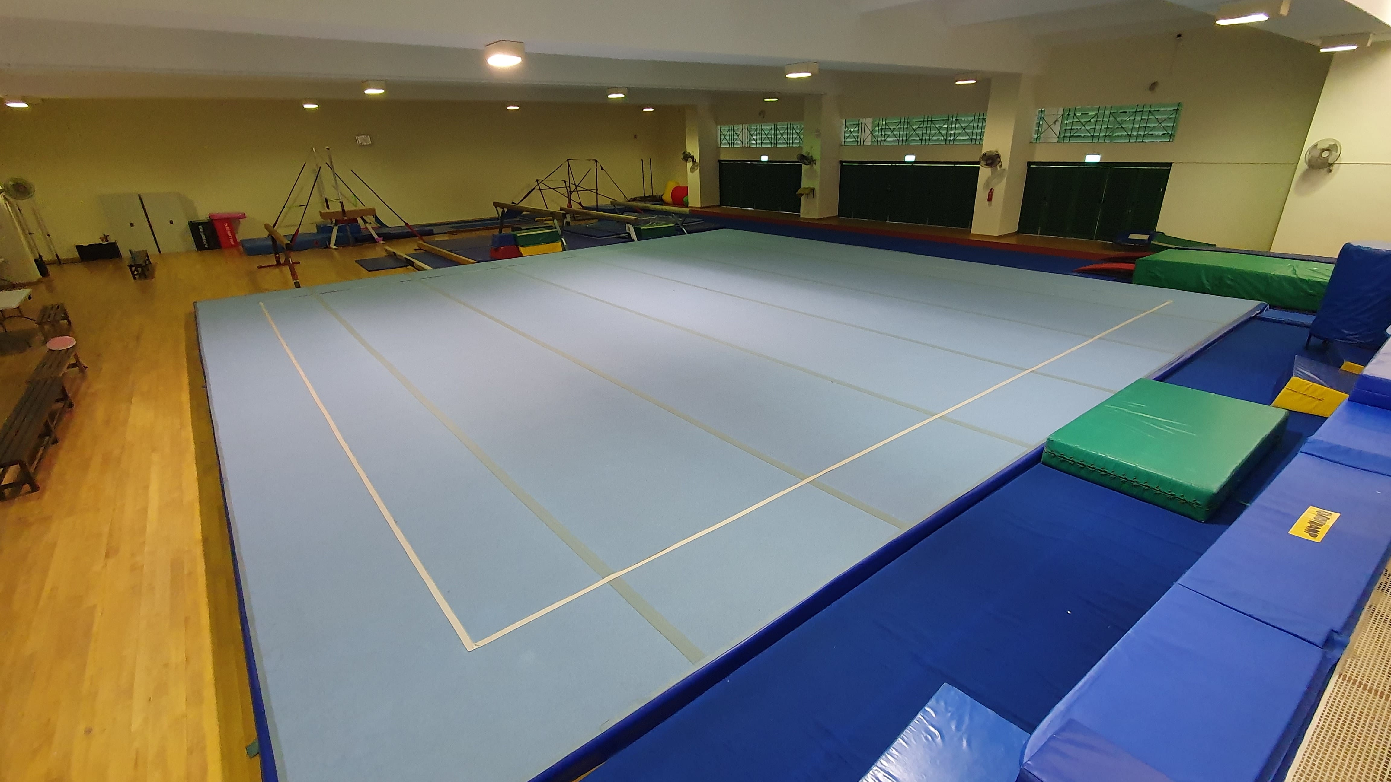 Indoor gymnastics gym with a large blue floor mat, blue padding around edges, and various apparatus in the background.