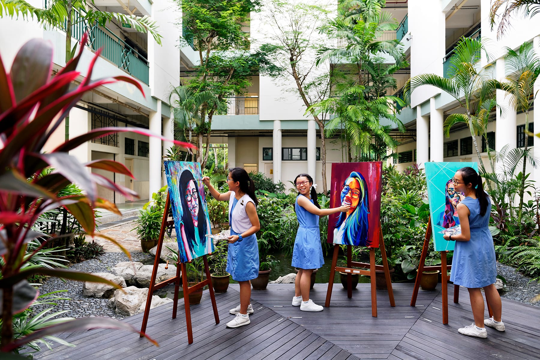 Three girls in blue uniforms paint colorful portrait canvases on easels in a lush outdoor school courtyard.