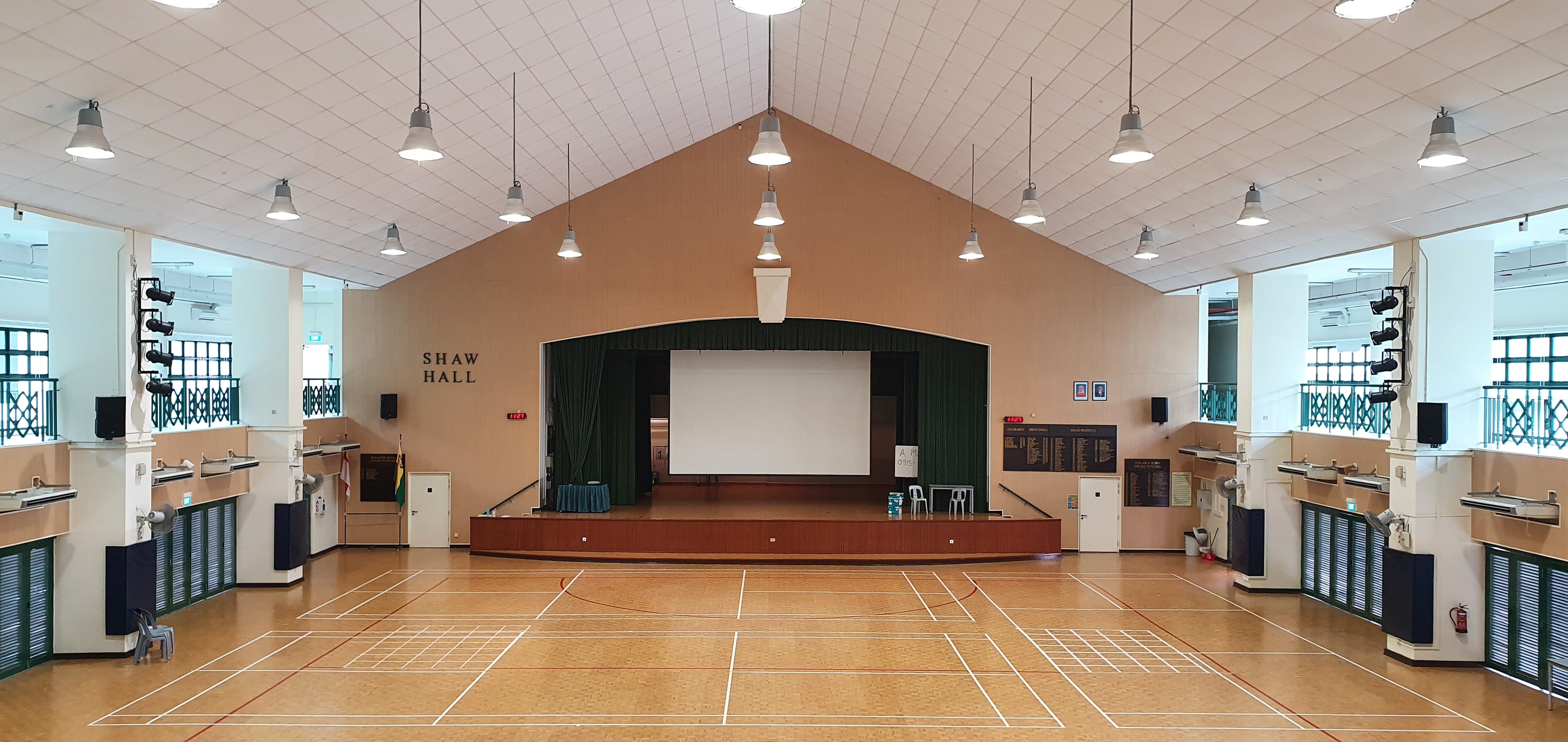 Empty Shaw Hall interior with a raised stage, projection screen, green curtains, and a wood sports court floor.