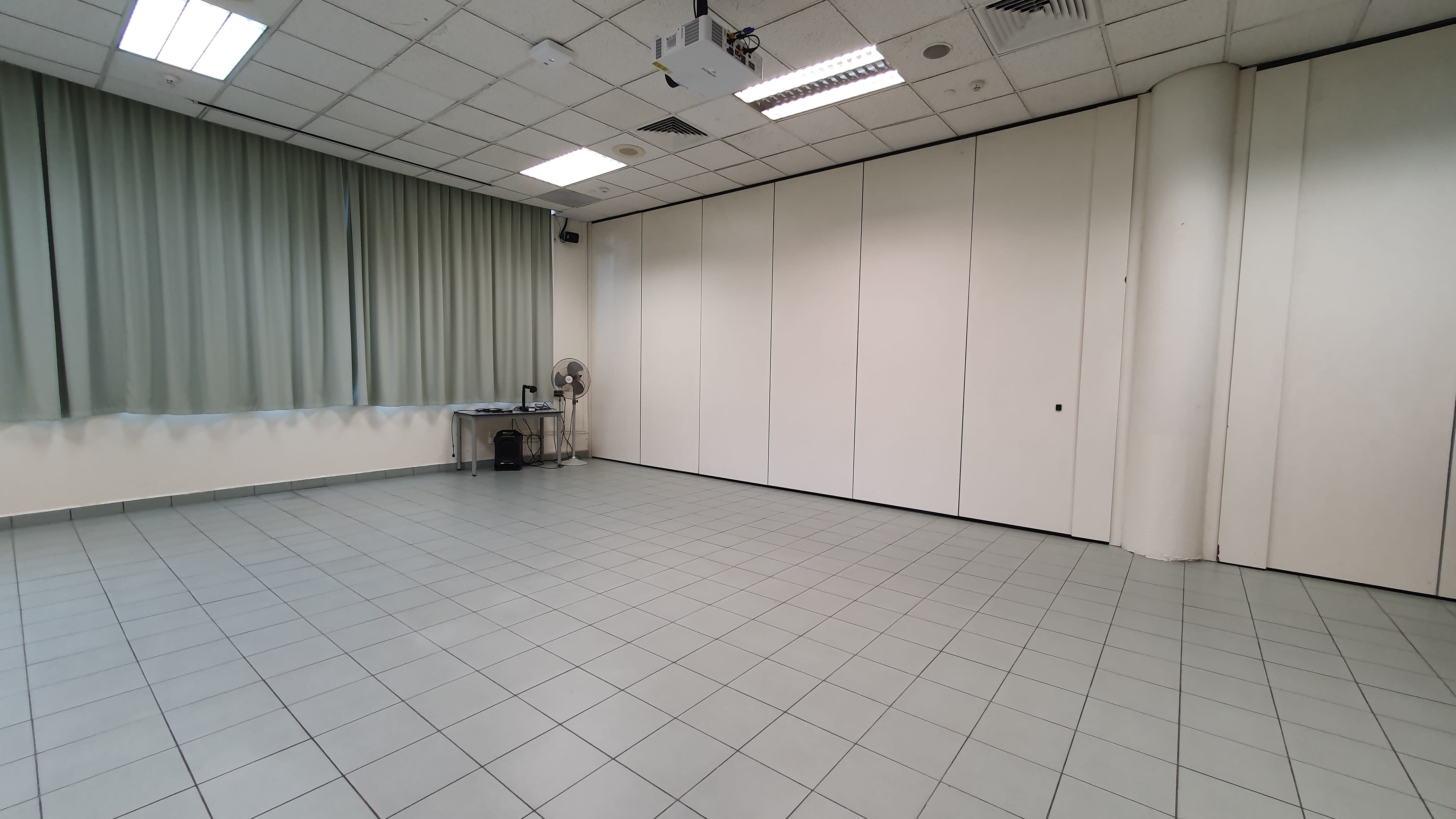 Empty room with white tile floor, ceiling projector, green curtains, folding partition walls, and a small desk with a fan.