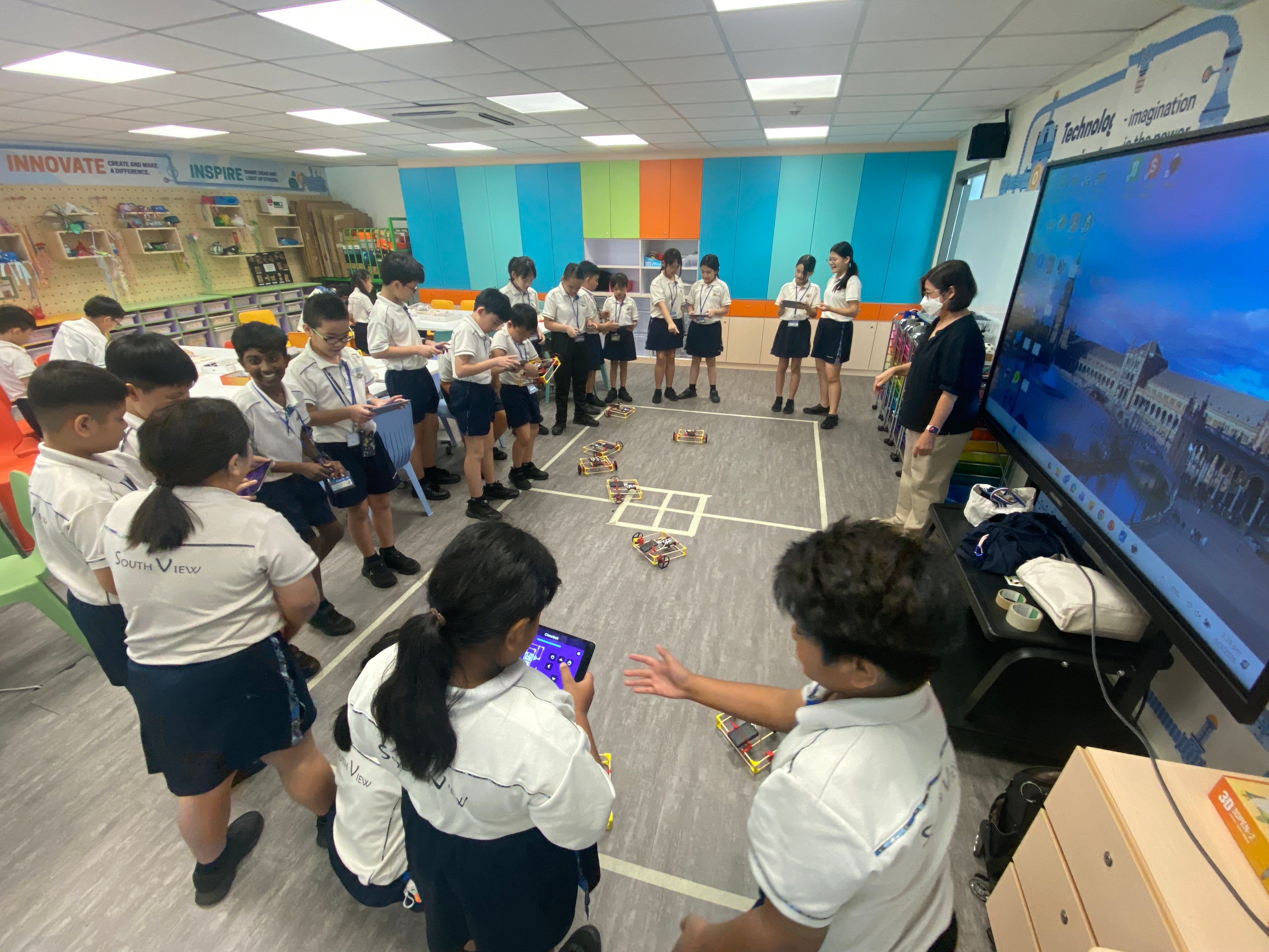 Students in uniforms surround small robots on a taped floor grid while a teacher watches in a classroom.