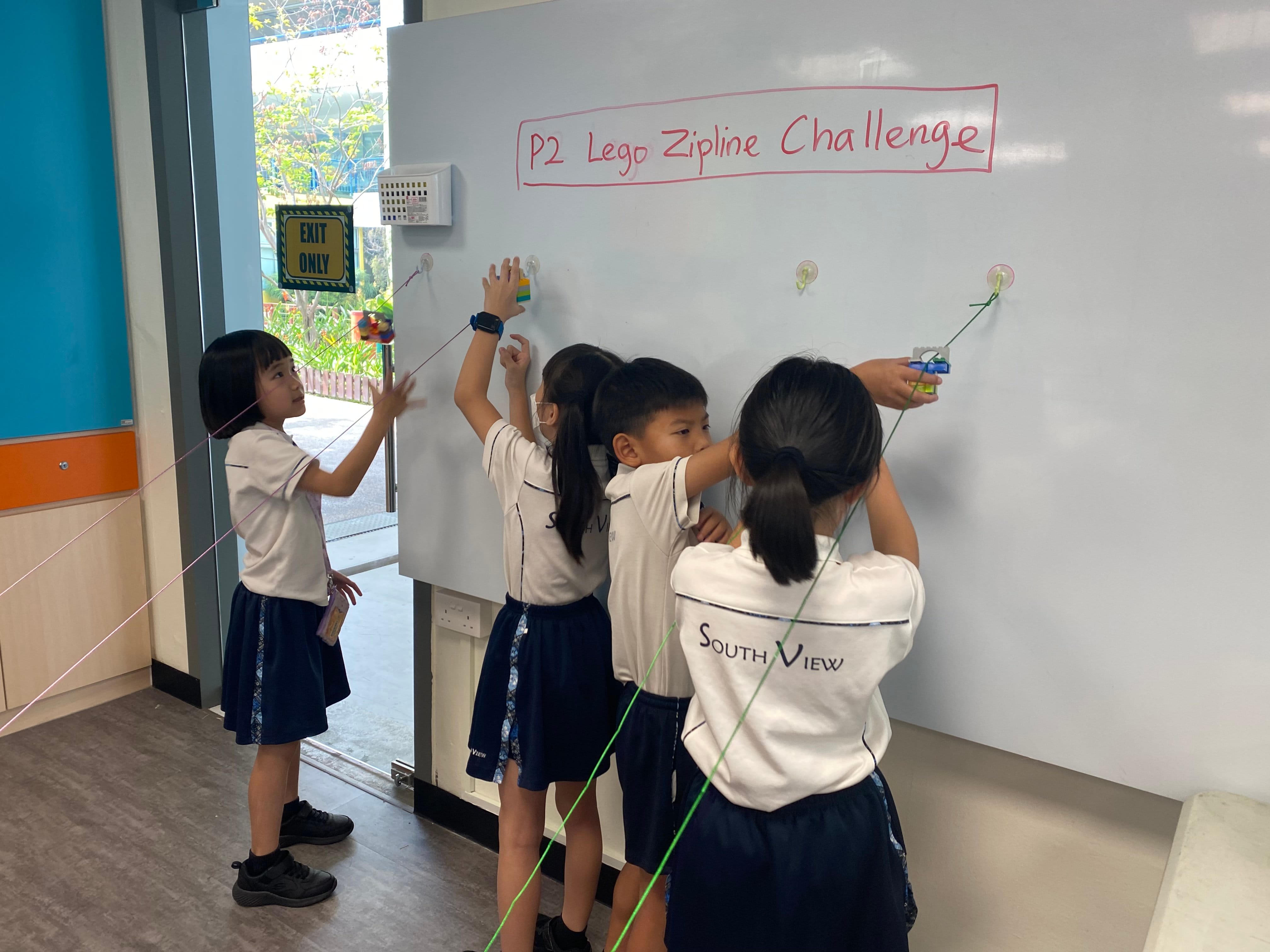 Four children in school uniforms attach Lego pieces to strings on a whiteboard labeled Lego Zipline Challenge