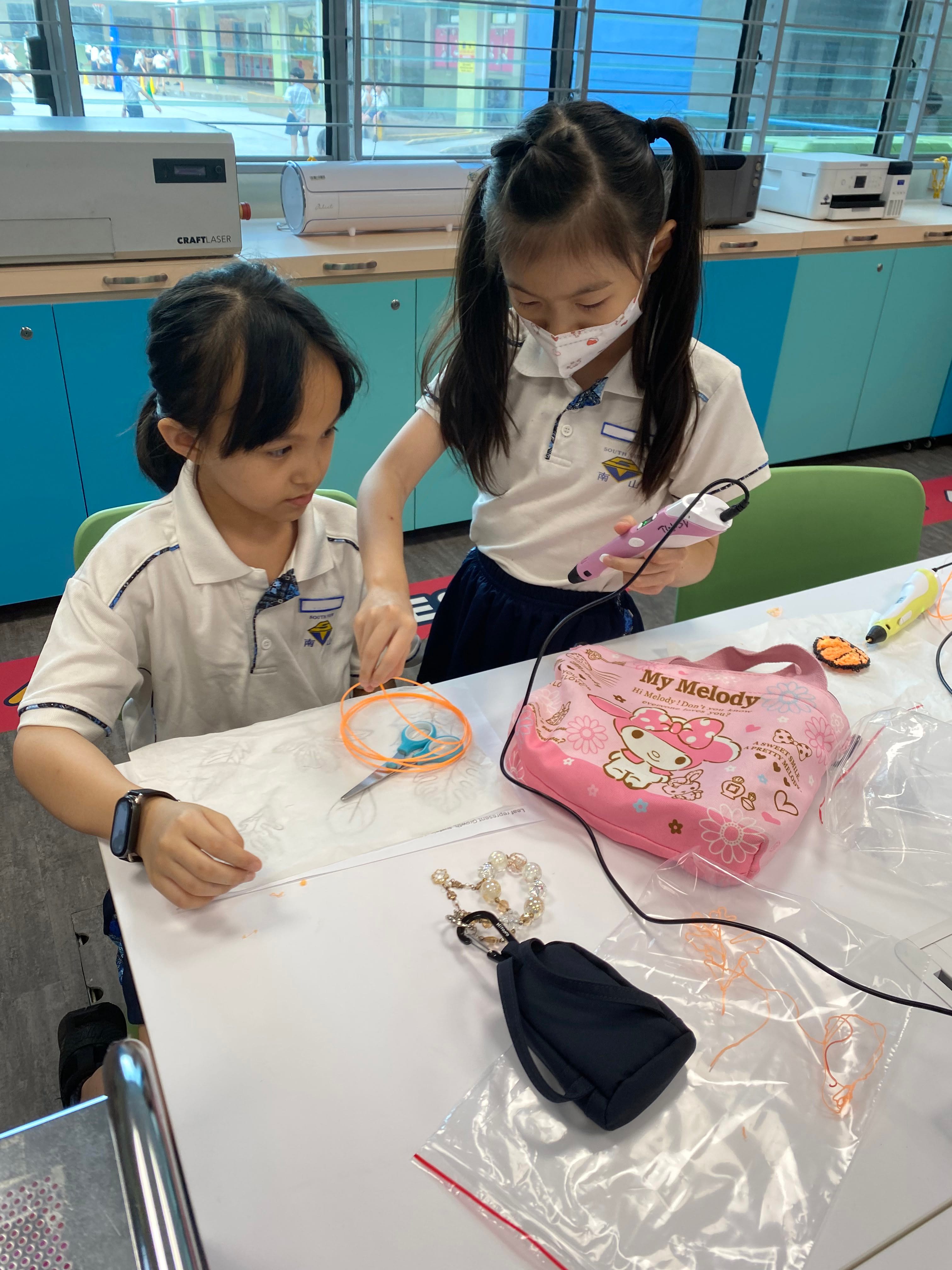 Two schoolgirls at a table, one using a purple 3D pen on orange filament beside a pink My Melody case.