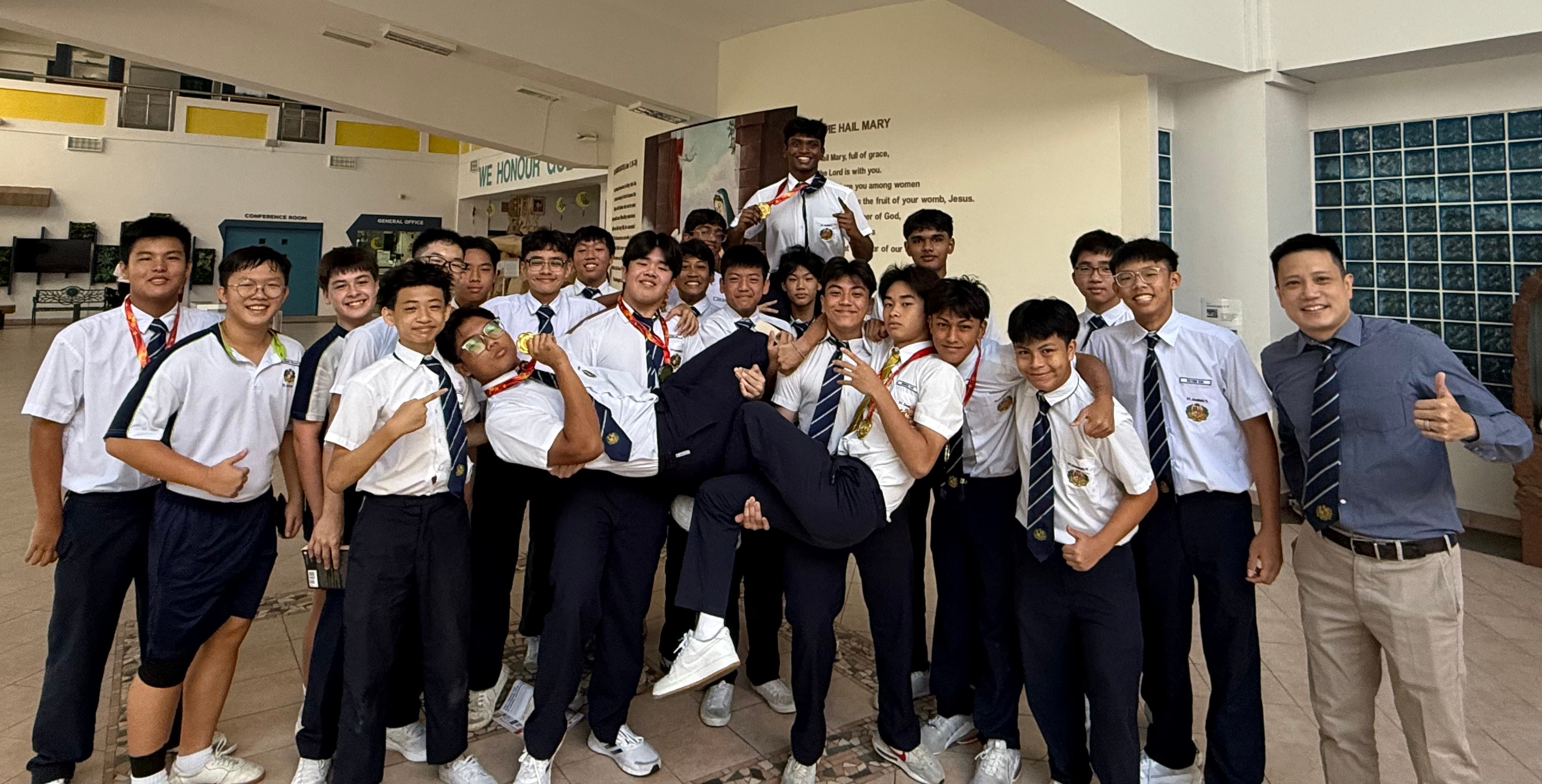 A large group of male students in white shirts and ties celebrate indoors, lifting one student horizontally while others show medals.