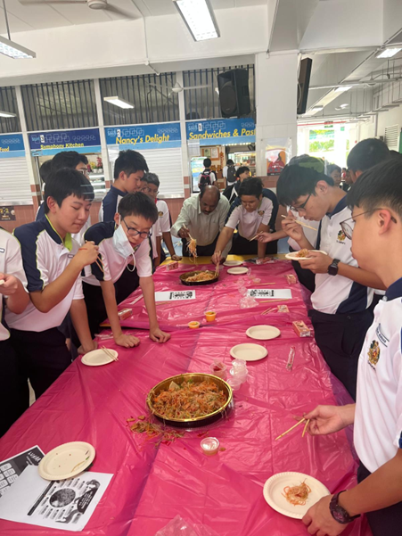 Students in uniforms gather around a pink-covered table eating food from large shared dishes with chopsticks.