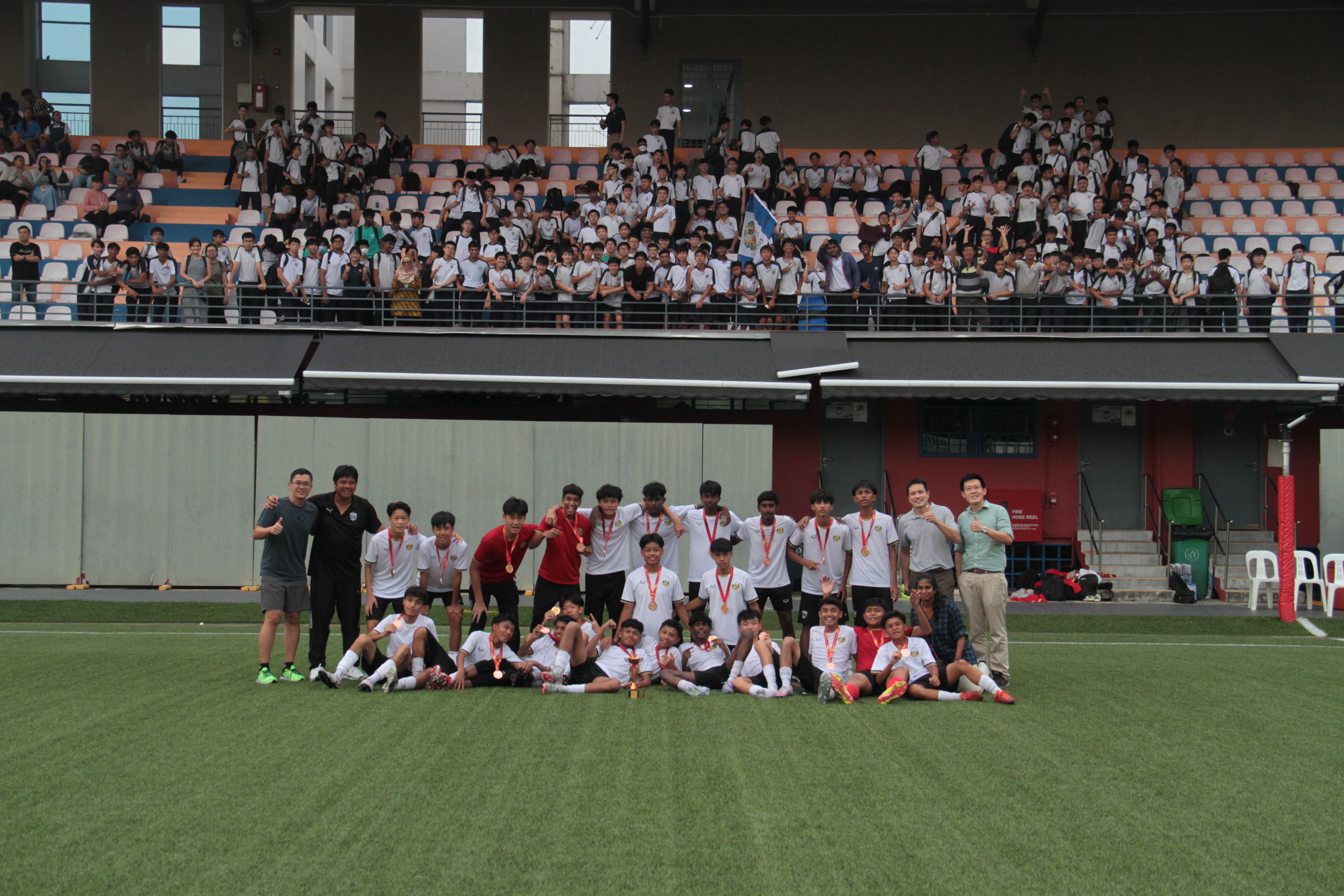 A youth football team wearing medals poses on a grass pitch with a large crowd of students in stadium stands behind them.