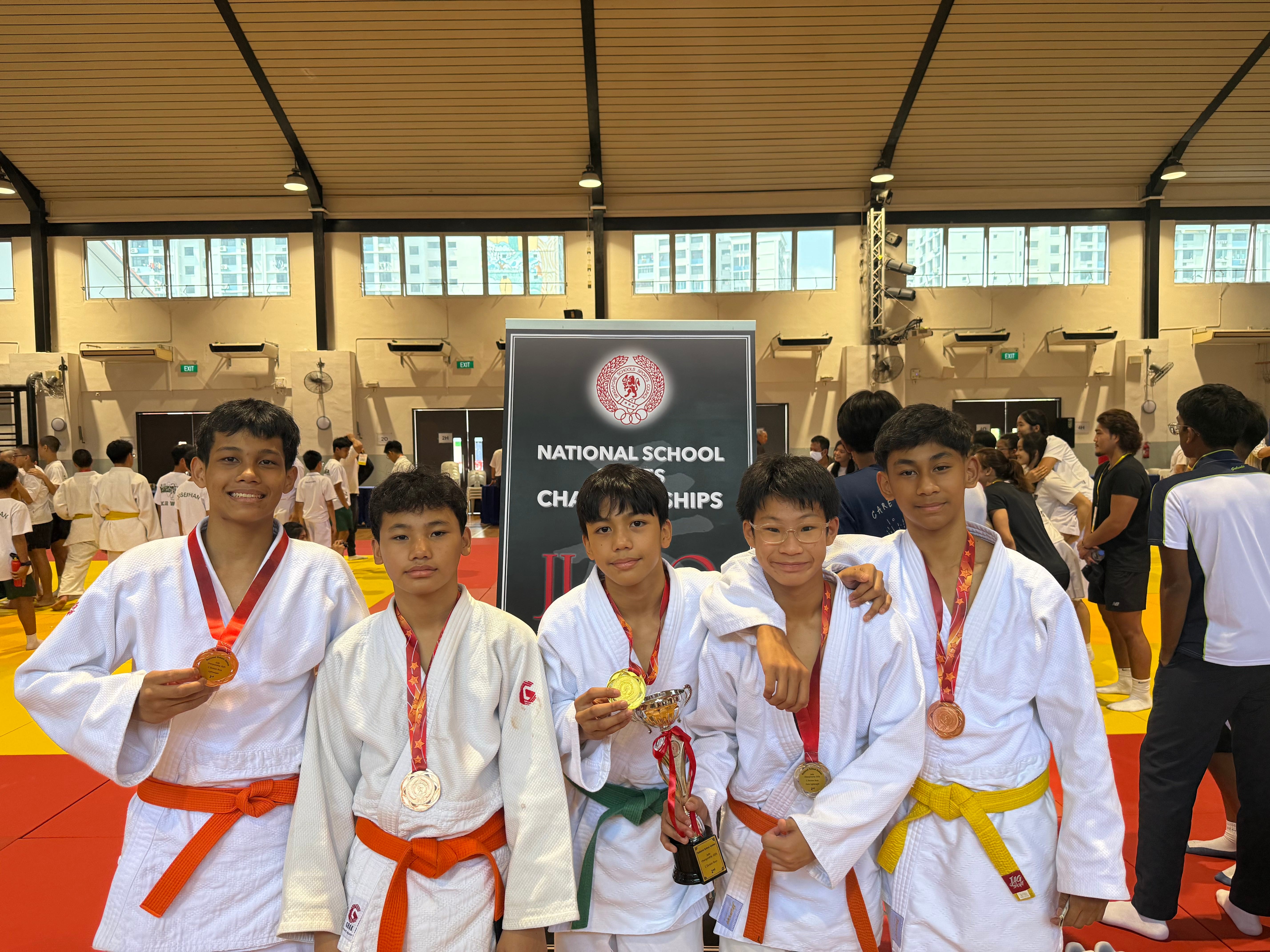 Five young judokas in white uniforms pose with medals and a trophy at a National School Championships event.