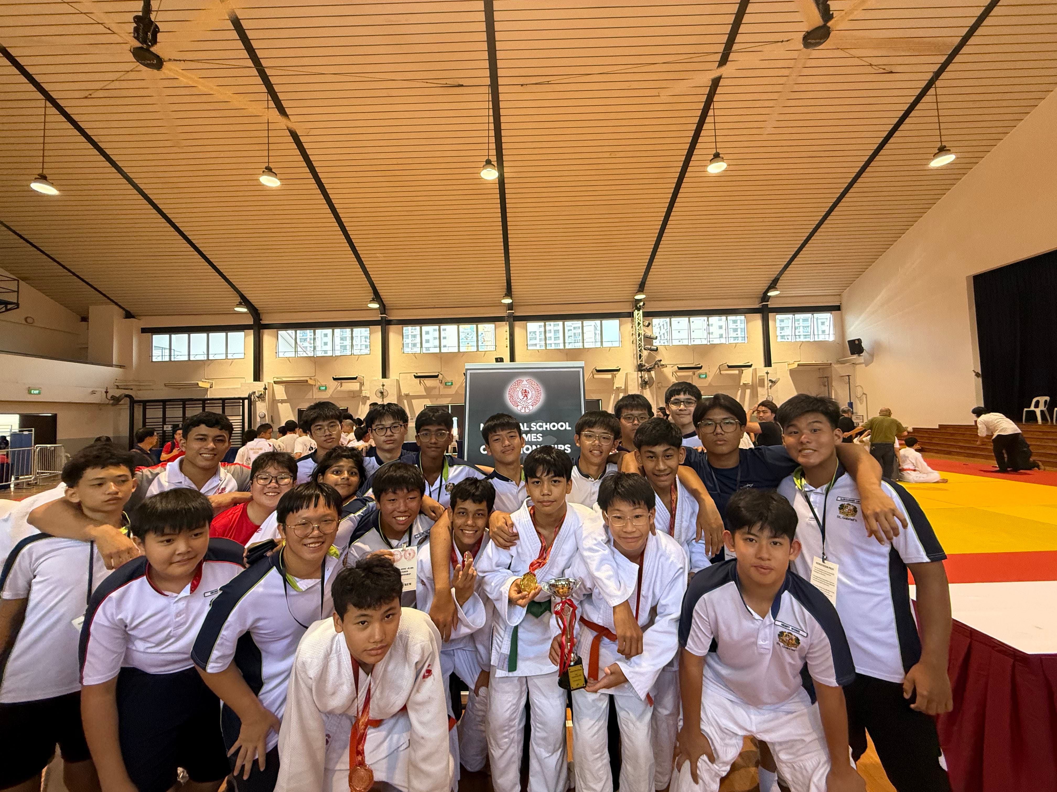 A large group of young male judo athletes posing together in a gym, holding a trophy and medals at a school championship.